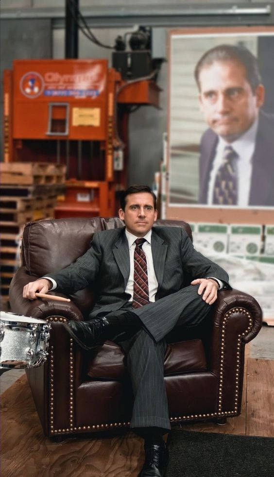 Michael Scott 桌布 壁紙 — 一個穿著細條紋西裝的男人自信地坐在一個倉庫環境中的棕色皮革扶手椅上，身後有一幅他自己的大型肖像。