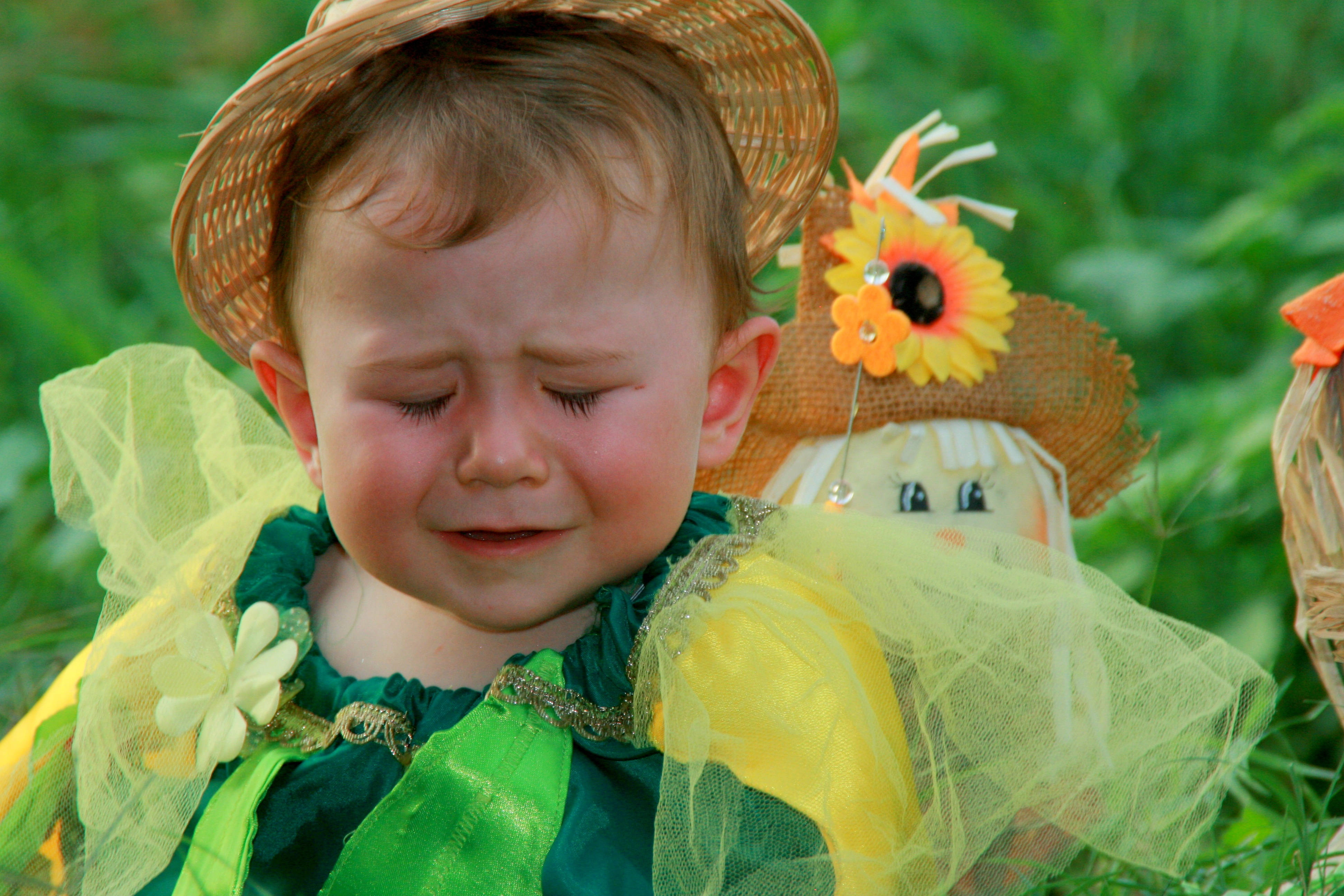 crying sad boy 桌布 壁紙 — 一個穿著綠色和黃色服裝、戴著草帽的哭泣幼兒坐在草地上稻草人旁邊。