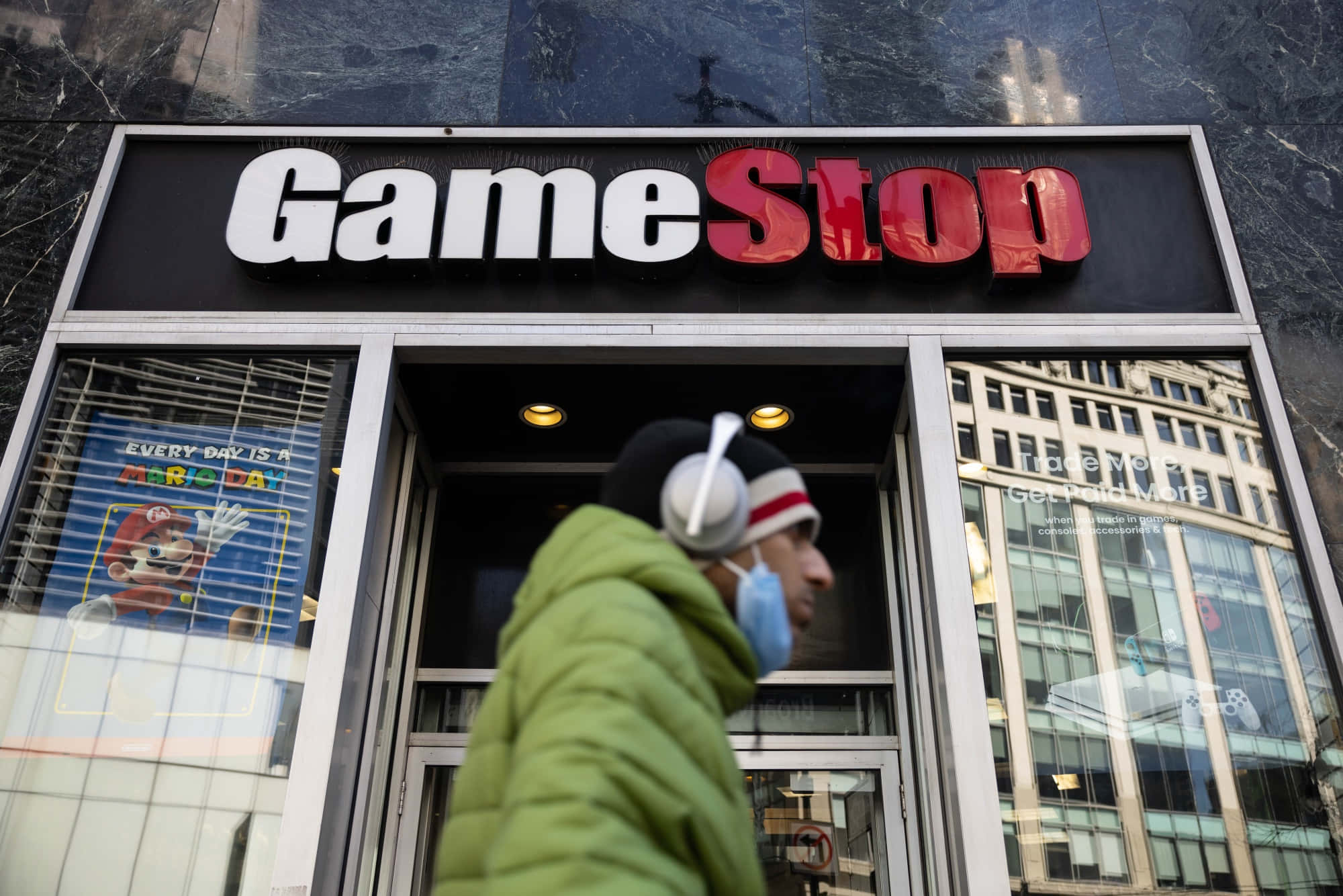 GameStop 桌布 壁紙 — 一個穿著綠色外套和戴耳機的人走過一間GameStop店，櫥窗裡有瑪利歐海報。