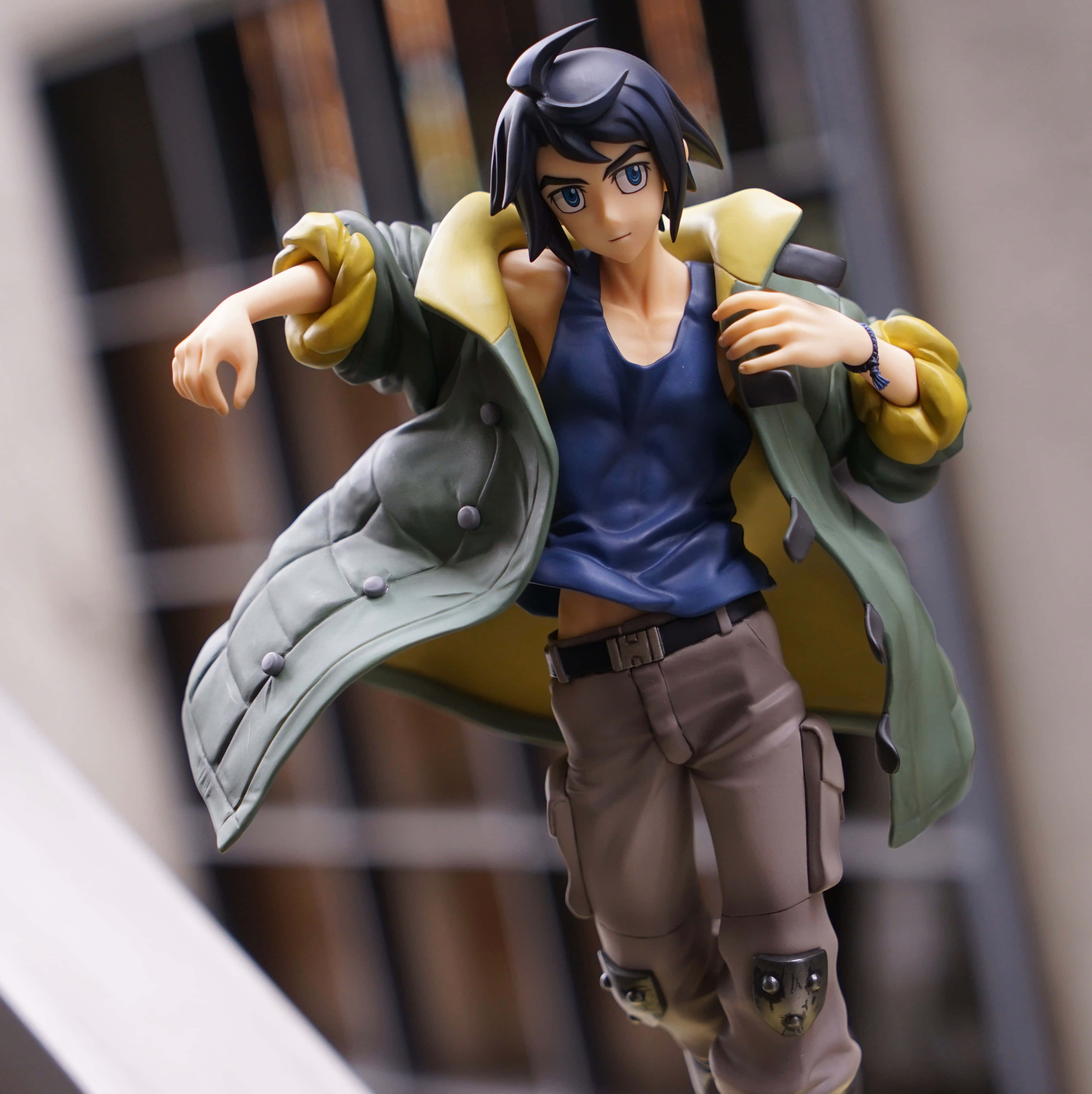 Mobile Suit Gundam Iron-blooded Orphans 桌布 壁紙 — 一個穿著綠色外套和藍色背心的年輕男性角色的詳細動漫人偶，在模糊的城市背景下動態擺姿勢。