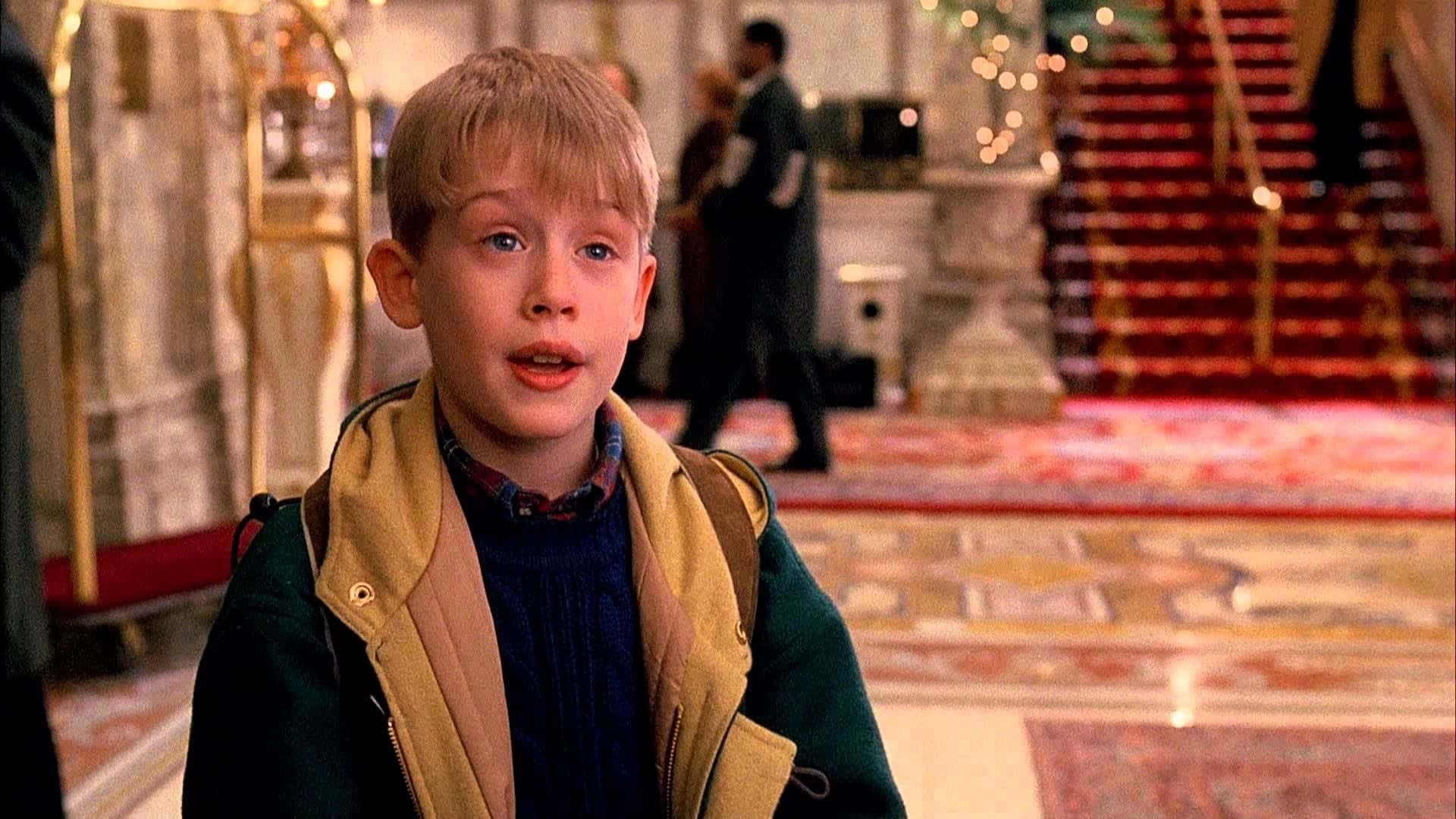 Funny Home Alone 桌布 壁紙 — 一個穿著綠色外套的小男孩站在一個鋪有紅色地毯樓梯和華麗裝飾的豪華飯店大廳。