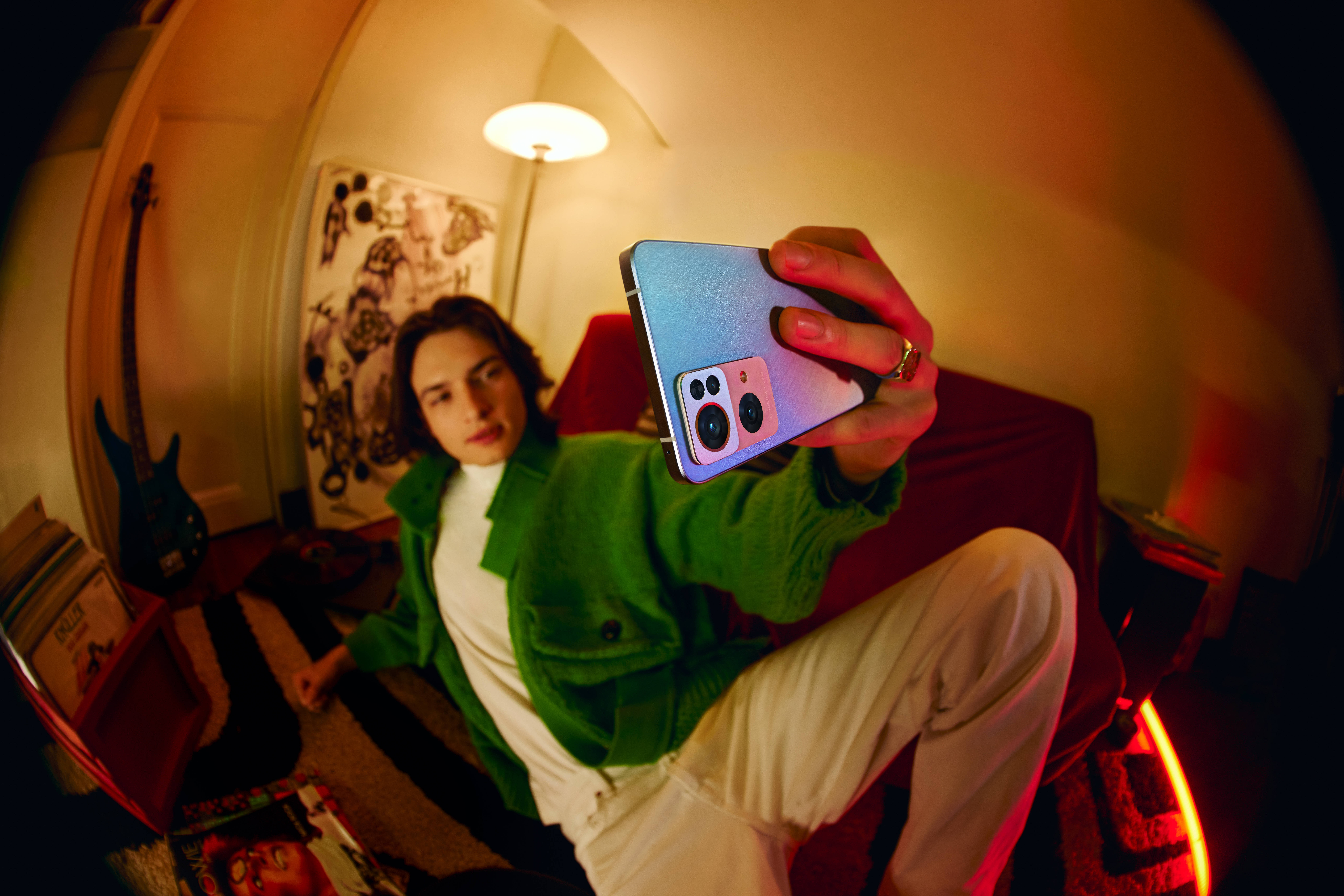 Oppo 桌布 壁紙 — 一個穿著綠色外套的年輕人在一個溫暖光線、雜亂的房間裡用彩色手機拍攝魚眼自拍。