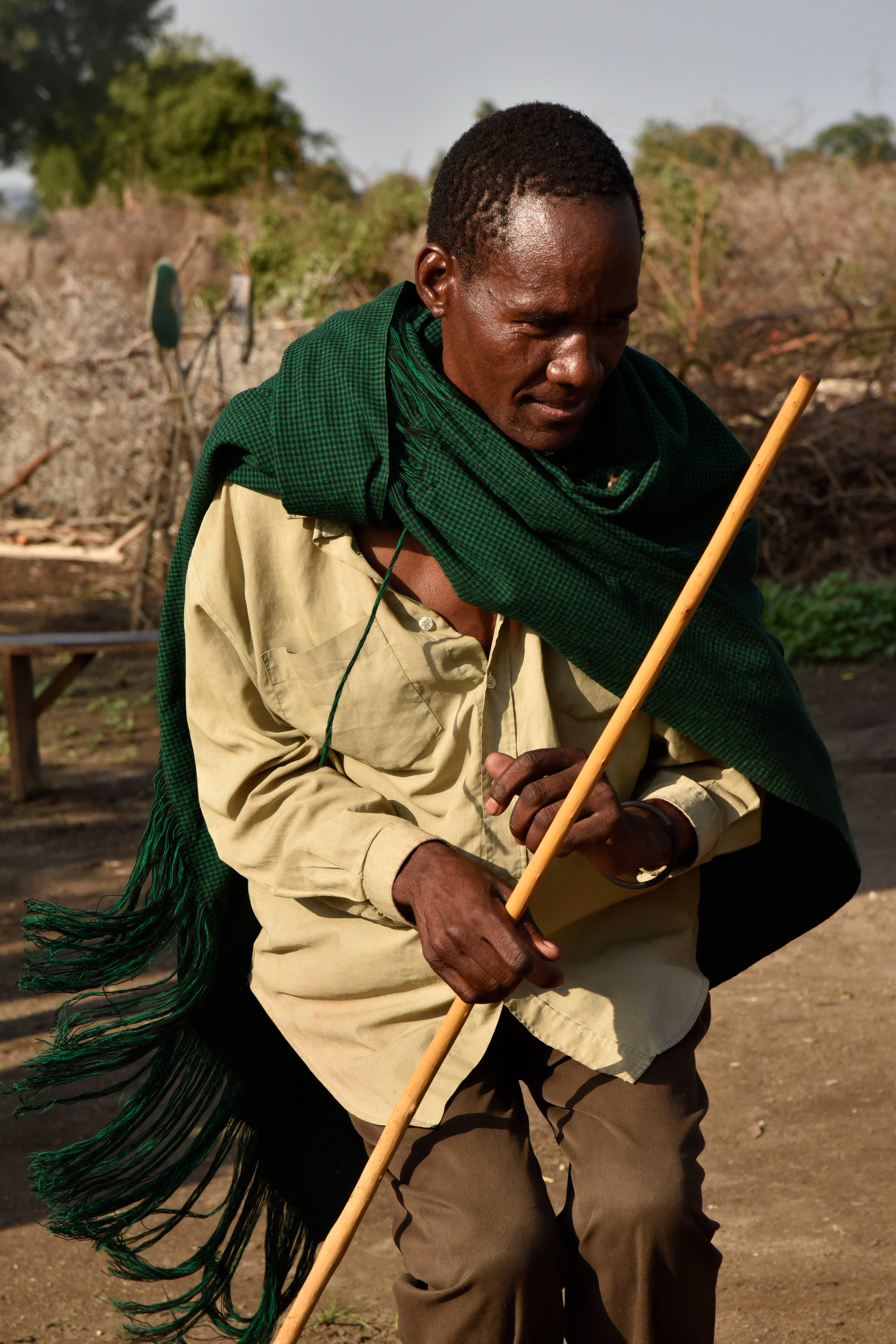 Tanzania 桌布 壁紙 — 一個穿著綠色披肩和米色襯衫的男人，在乾燥的戶外環境中跪著抓住一根木棍。