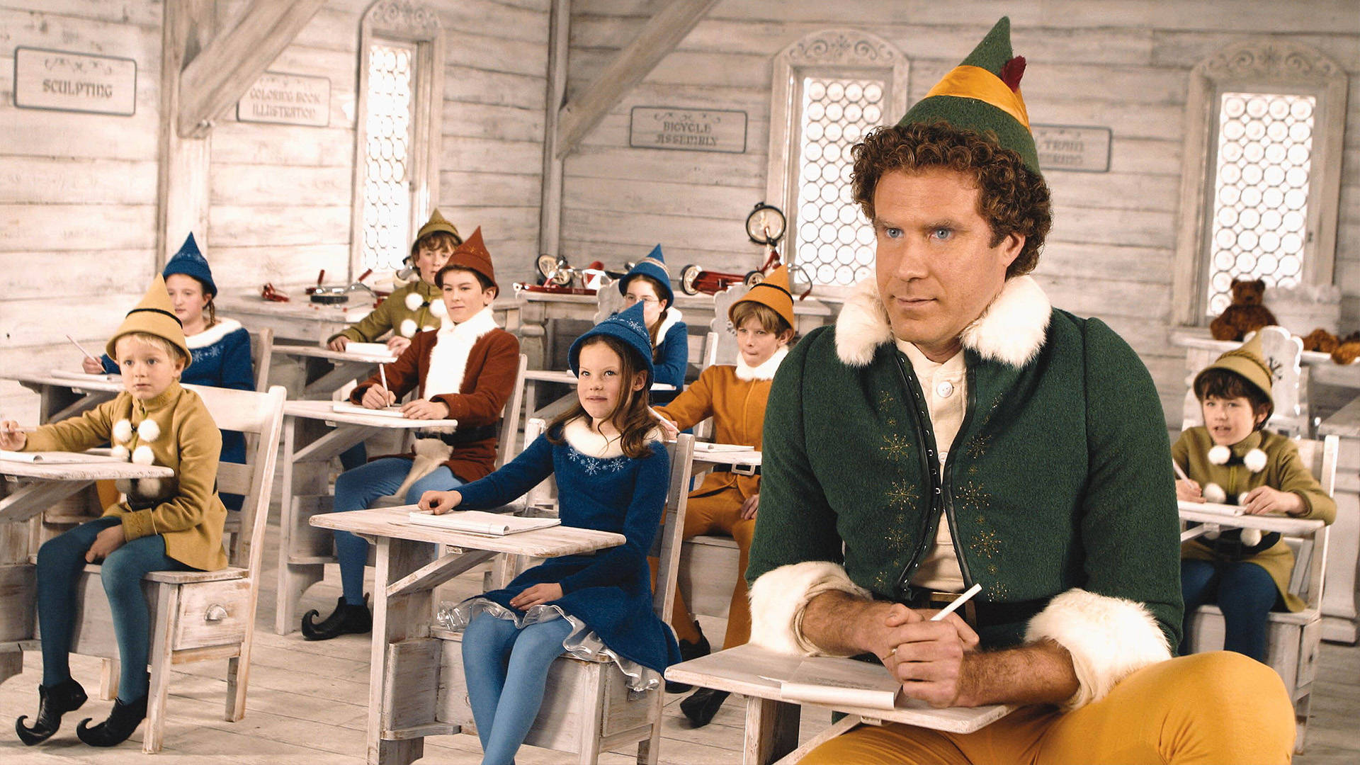 Elf Movie 桌布 壁紙 — 一個穿著綠色精靈服裝的男人坐在木製教室的書桌前，孩子們穿著精靈服裝在他們的書桌上寫字。