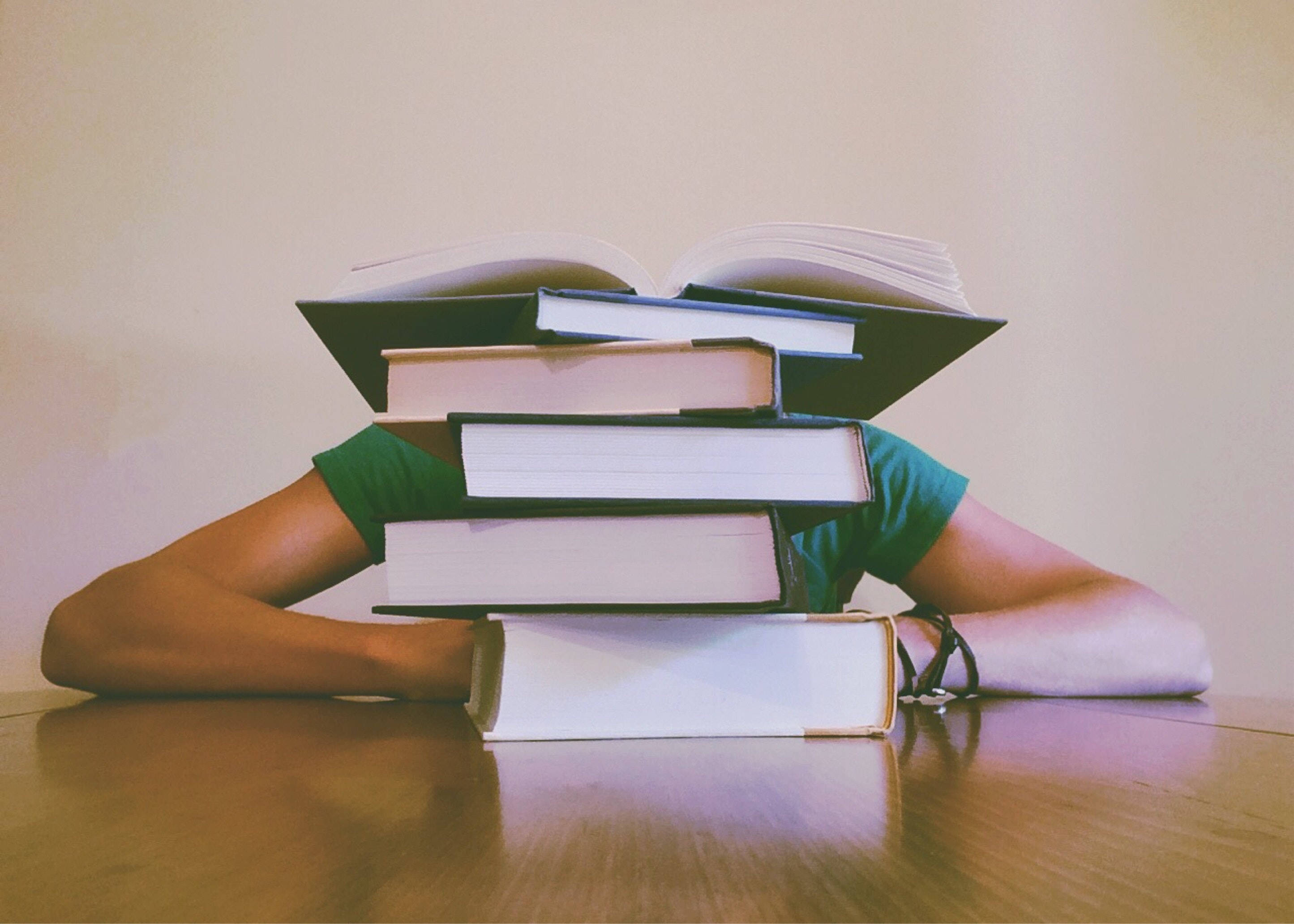 讀書動力 桌布 壁紙 — 一個穿著綠色襯衫的人將頭靠在木桌上高高堆起的書本下。