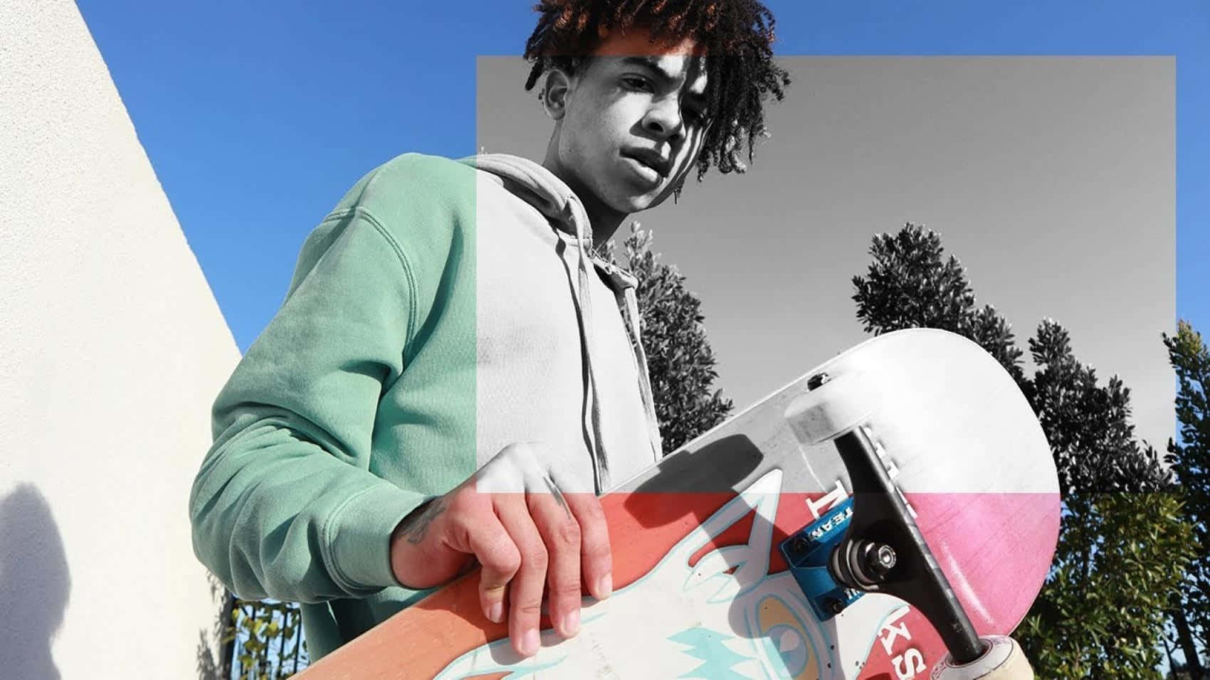 Hip Hop 桌布 壁紙 — 一個穿著綠色連帽衫的年輕滑板手，拿著色彩繽紛的滑板，背景是分開的藍天和單色樹木。