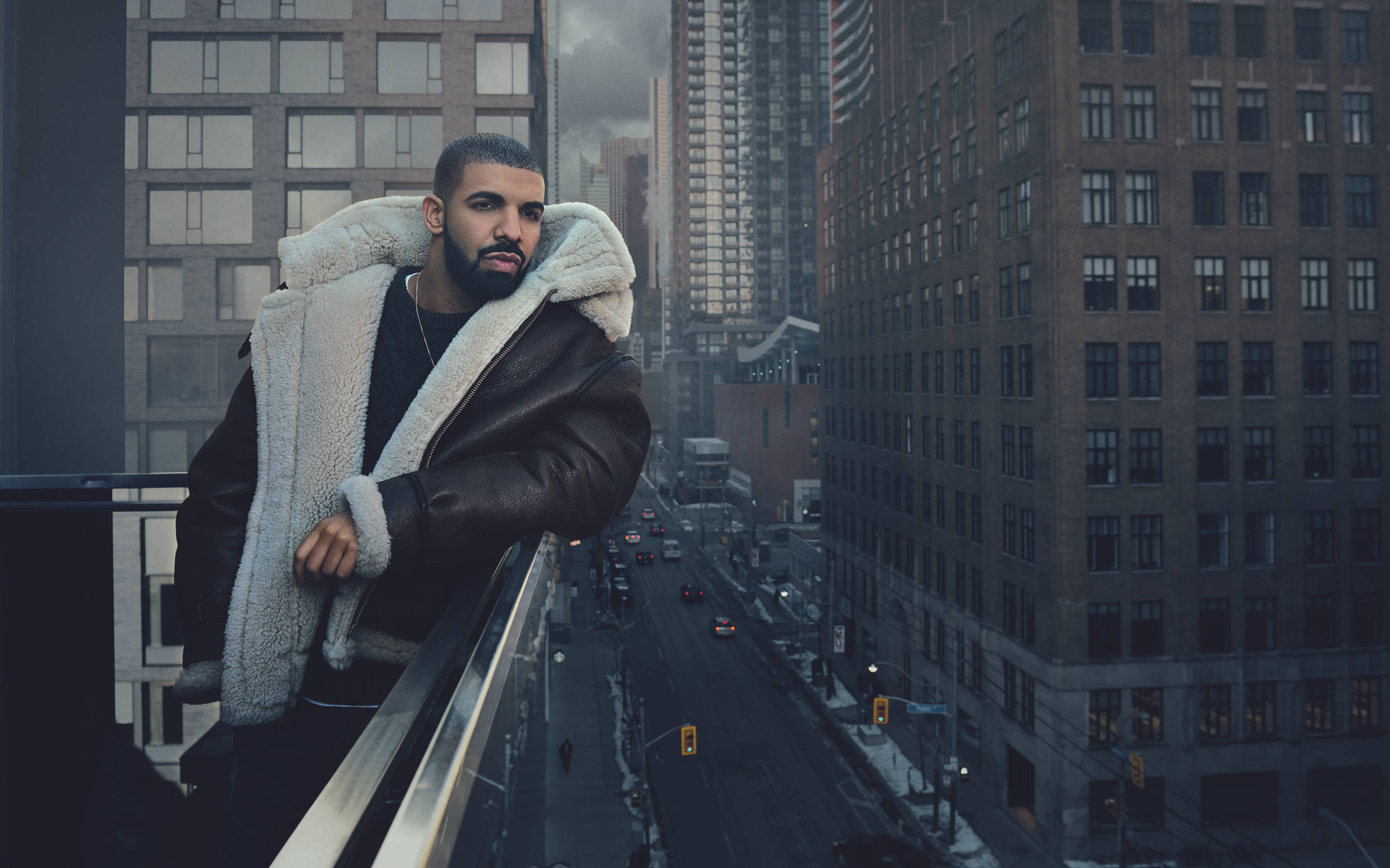 Drake 桌布 壁紙 — 一個穿著羊皮襯裡夾克的男人靠在陽台上，俯瞰高樓之間積雪的城市街道。