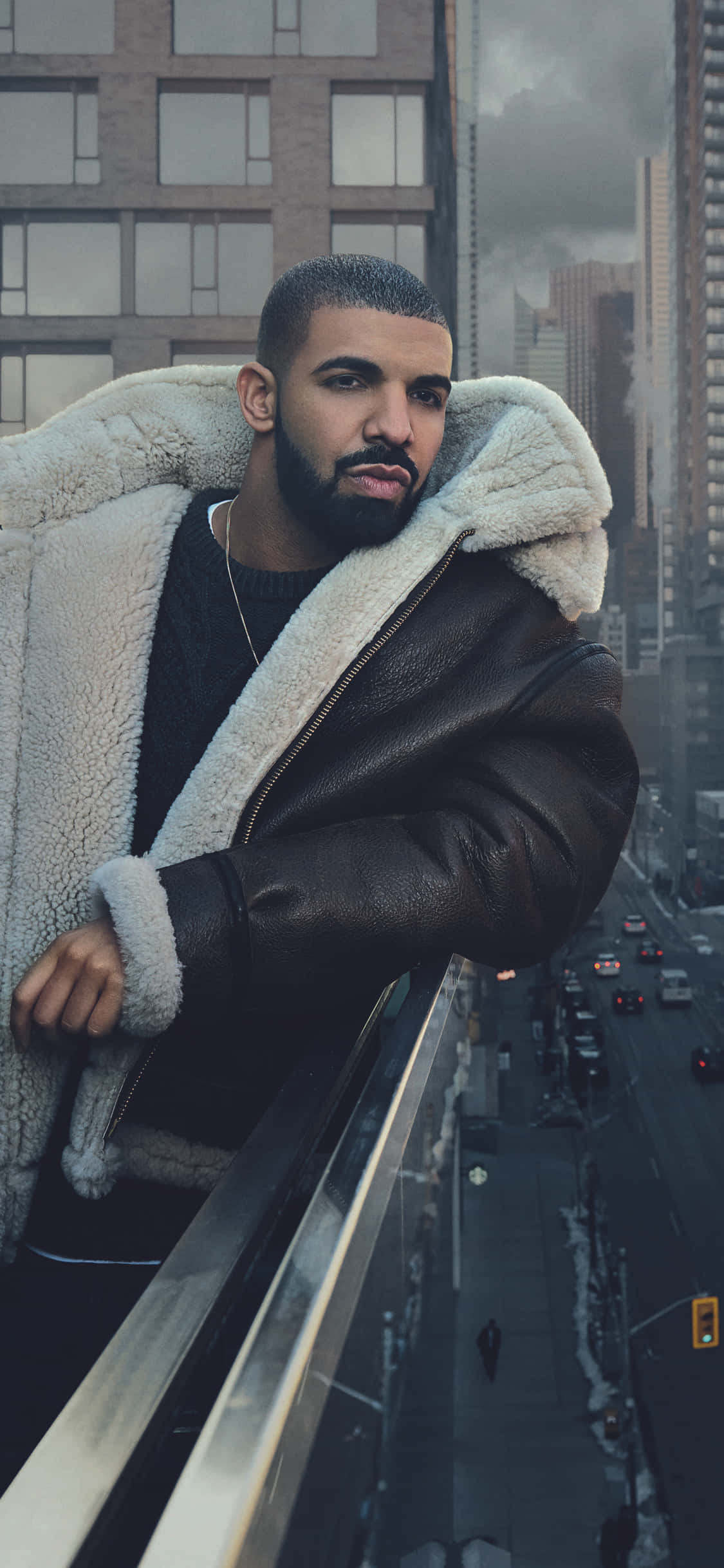Aesthetic Drake 桌布 壁紙 — 一個穿著羊羔毛內裡皮夾克的男人倚在高層建築的陽台上，俯瞰下方繁忙的城市街道。