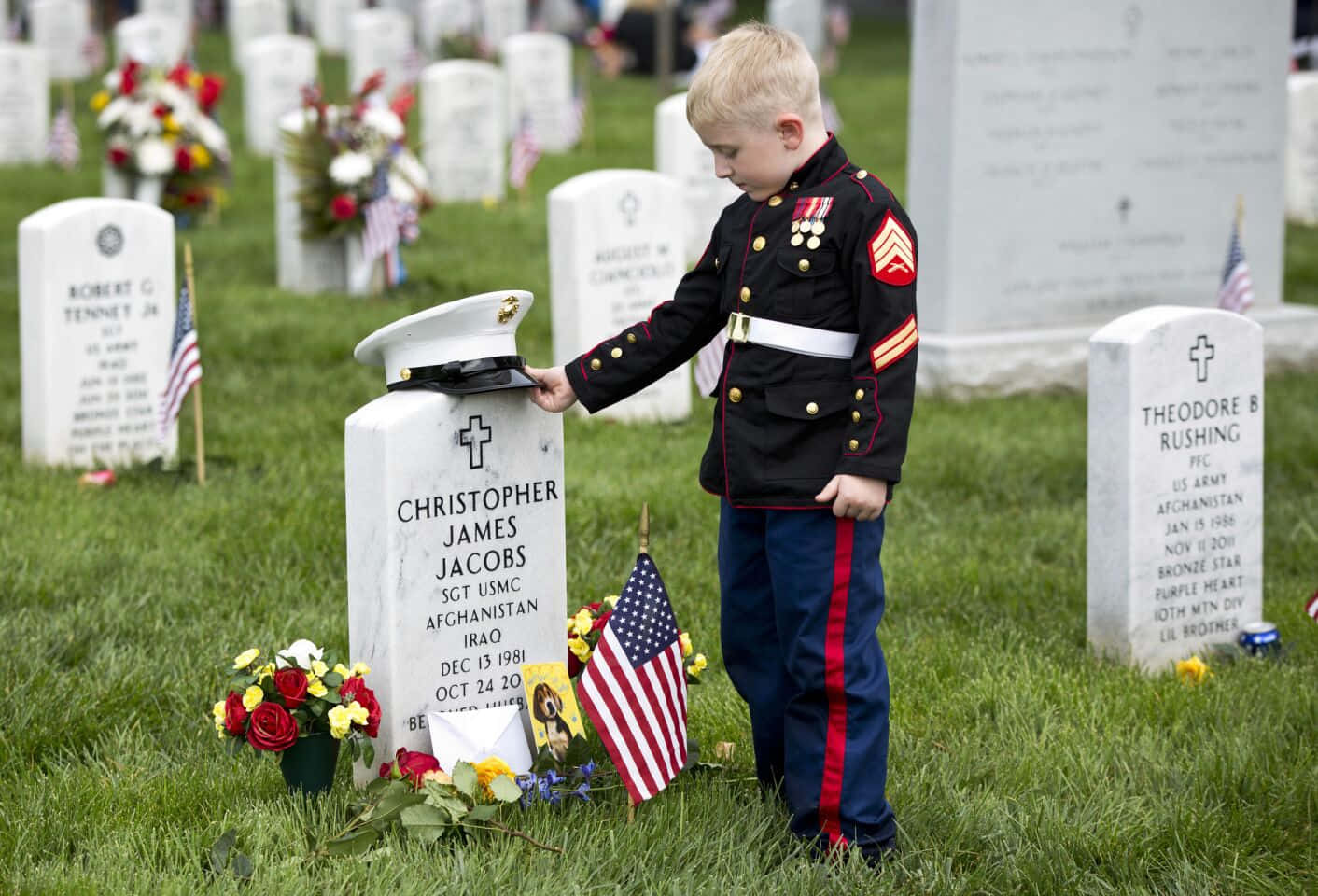 Memorial Day 桌布 壁紙 — 一個穿著美國海軍陸戰隊制服的小男孩在一個有美國國旗和鮮花的軍事墓地中，將帽子放在墓碑上。