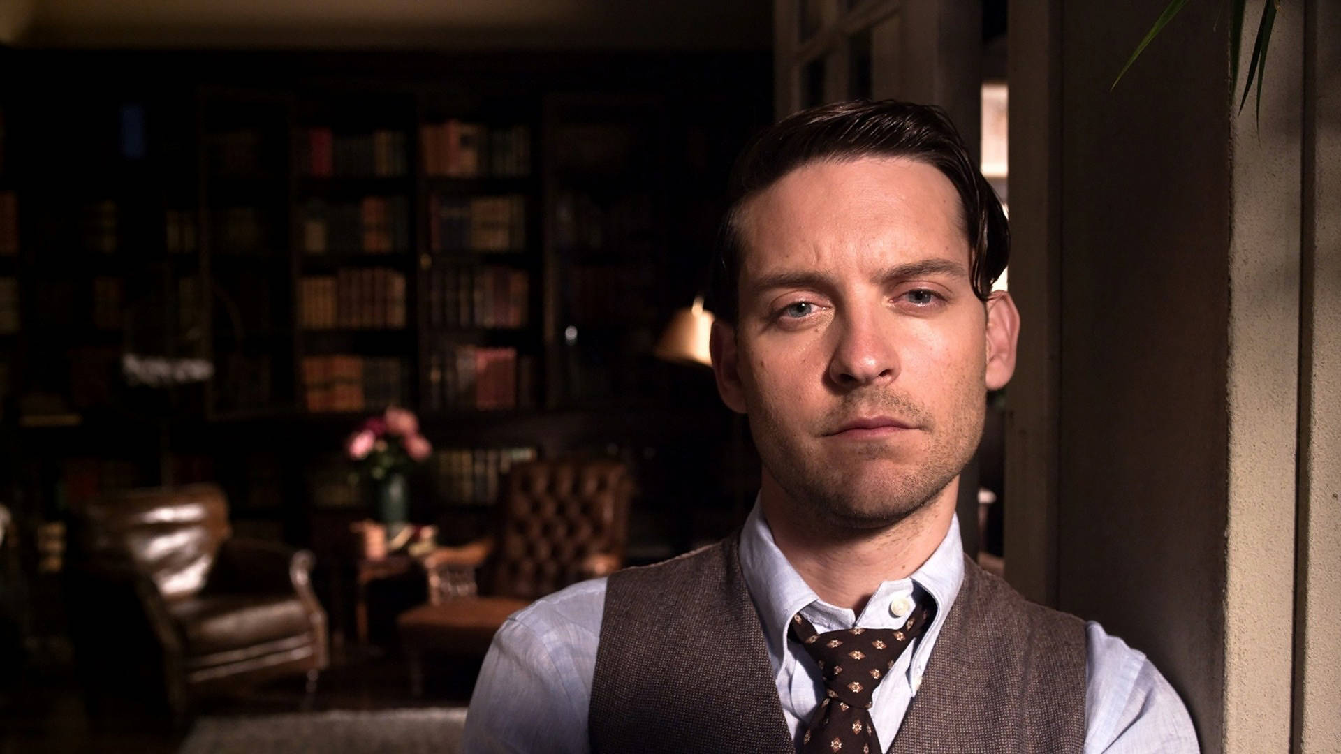 Tobey Maguire 桌布 壁紙 — 一個穿著背心和領帶的男人站在燈光昏暗的圖書館裡，身後有書架和皮椅。