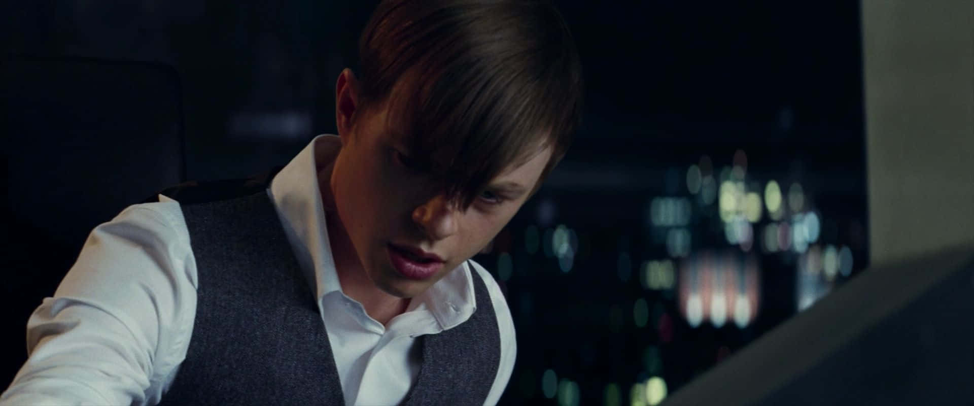 Norman Osborn 桌布 壁紙 — 一個穿著背心的年輕男子在昏暗的房間裡專注地向前傾，身後是模糊的城市燈光。