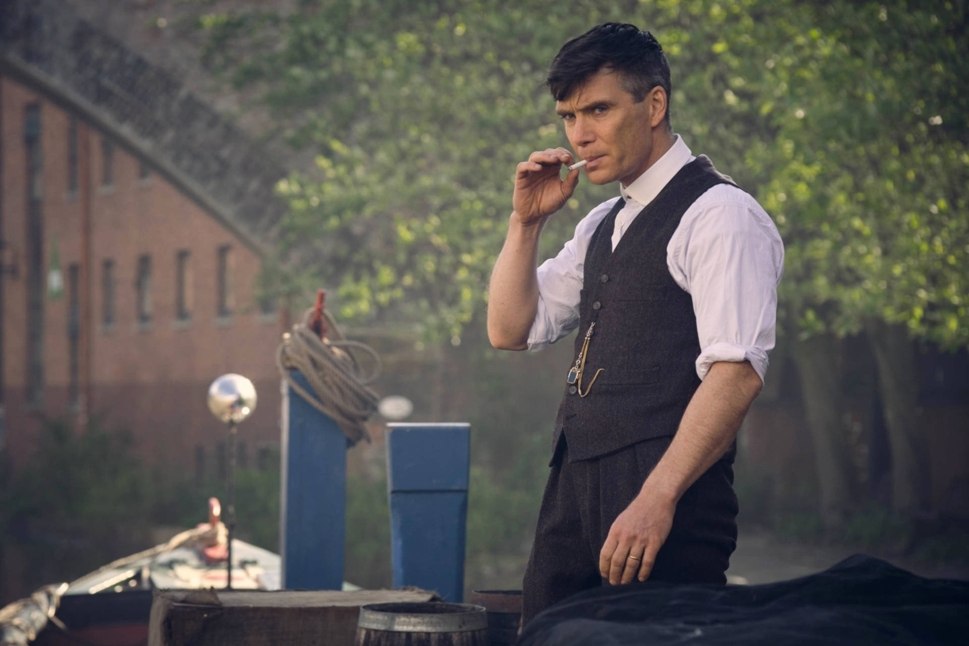Thomas Shelby 桌布 壁紙 — 一個穿著背心的男人在碼頭上，旁邊有一艘船，背景有磚建築和綠樹，正在抽煙。