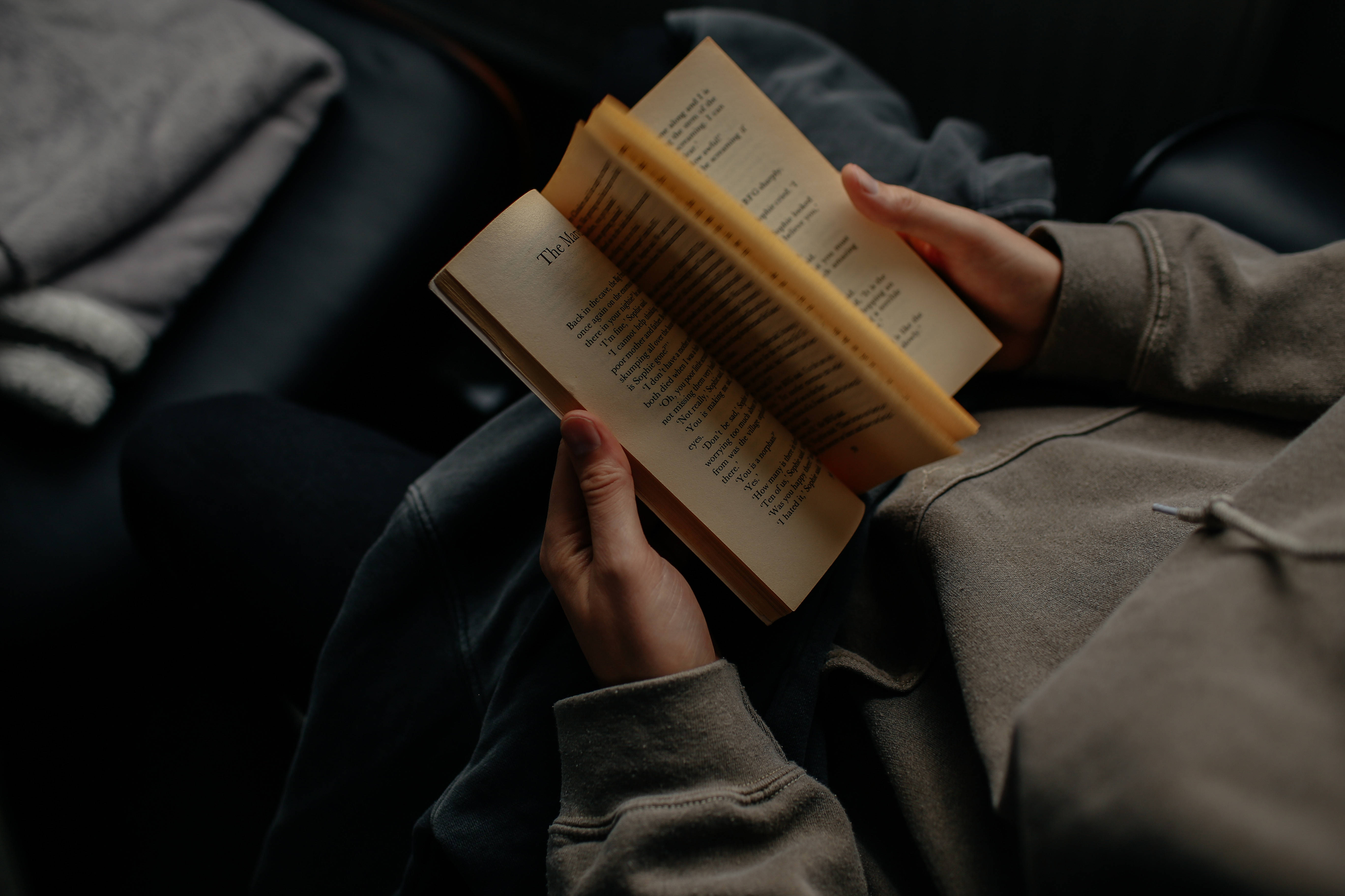 Reading 桌布 壁紙 — 一個穿著舒適連帽衫的人坐在深色沙發上，附近有柔軟的毯子，正在閱讀一本書。