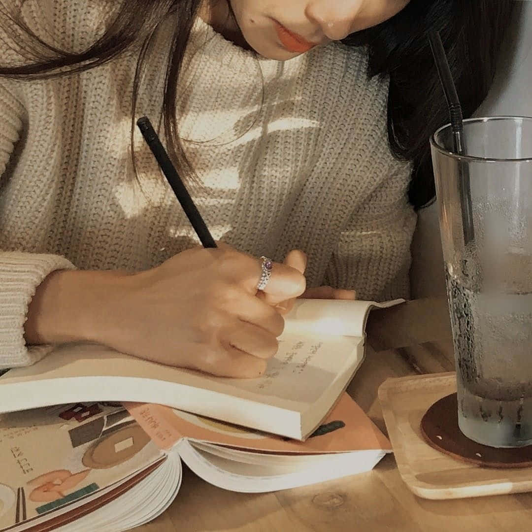 Study Aesthetic 桌布 壁紙 — 一個穿著舒適針織毛衣的人正在木桌上的一杯冰飲旁邊的筆記本上寫字。
