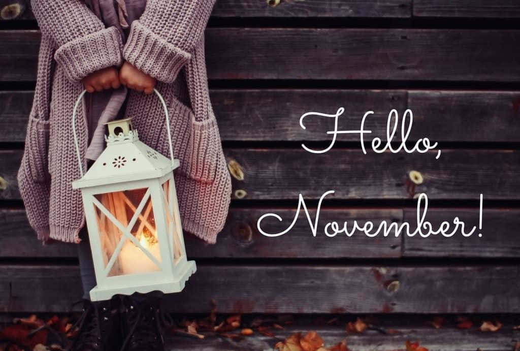 Cute November 桌布 壁紙 — 一個穿著舒適針織毛衣的孩子，拿著發光的燈籠，靠著一個帶有落葉和文字「你好十一月」的質樸木柵欄。