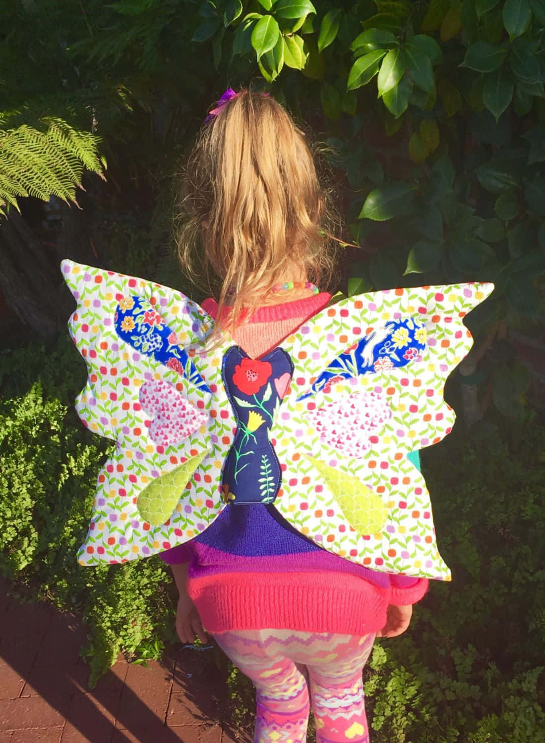Butterfly Wing Dress 桌布 壁紙 — 一個穿著色彩繽紛拼布蝴蝶服裝的小孩背對鏡頭站在陽光普照的花園裡。