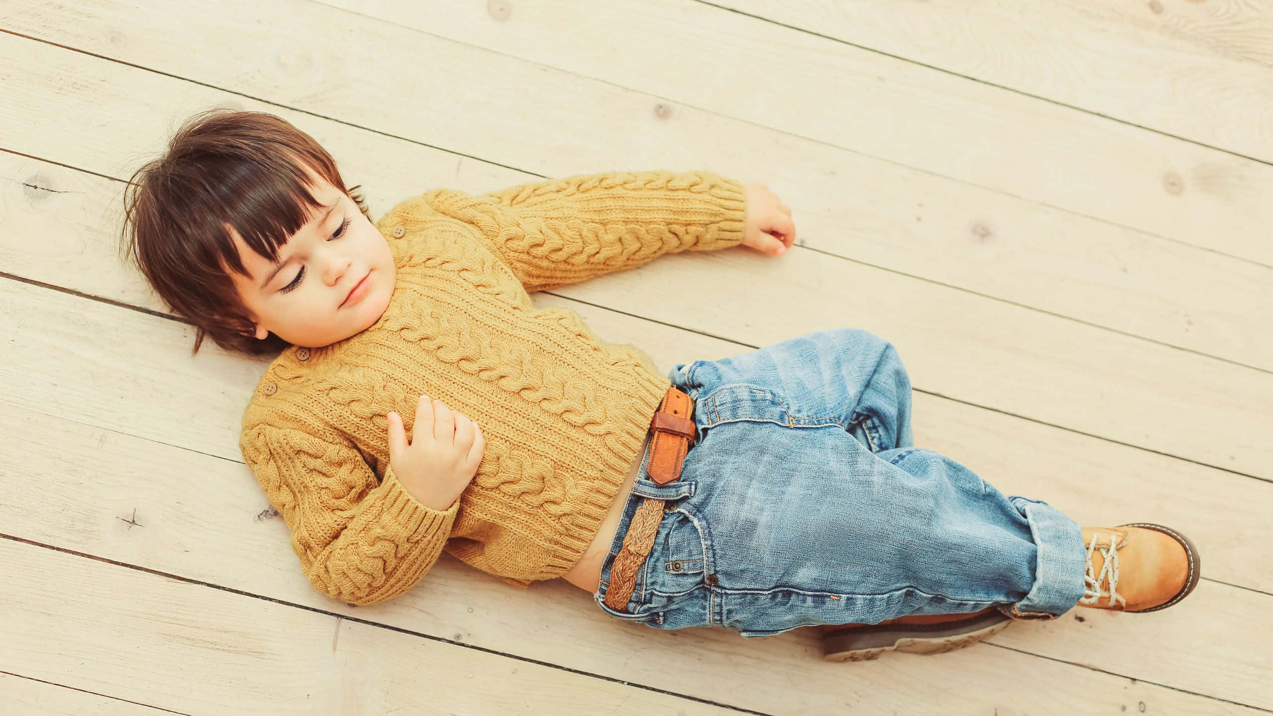 Cute Boy 桌布 壁紙 — 一個穿著芥末黃色麻花針織毛衣的睏倦幼兒，平靜地躺在淺色木地板上。