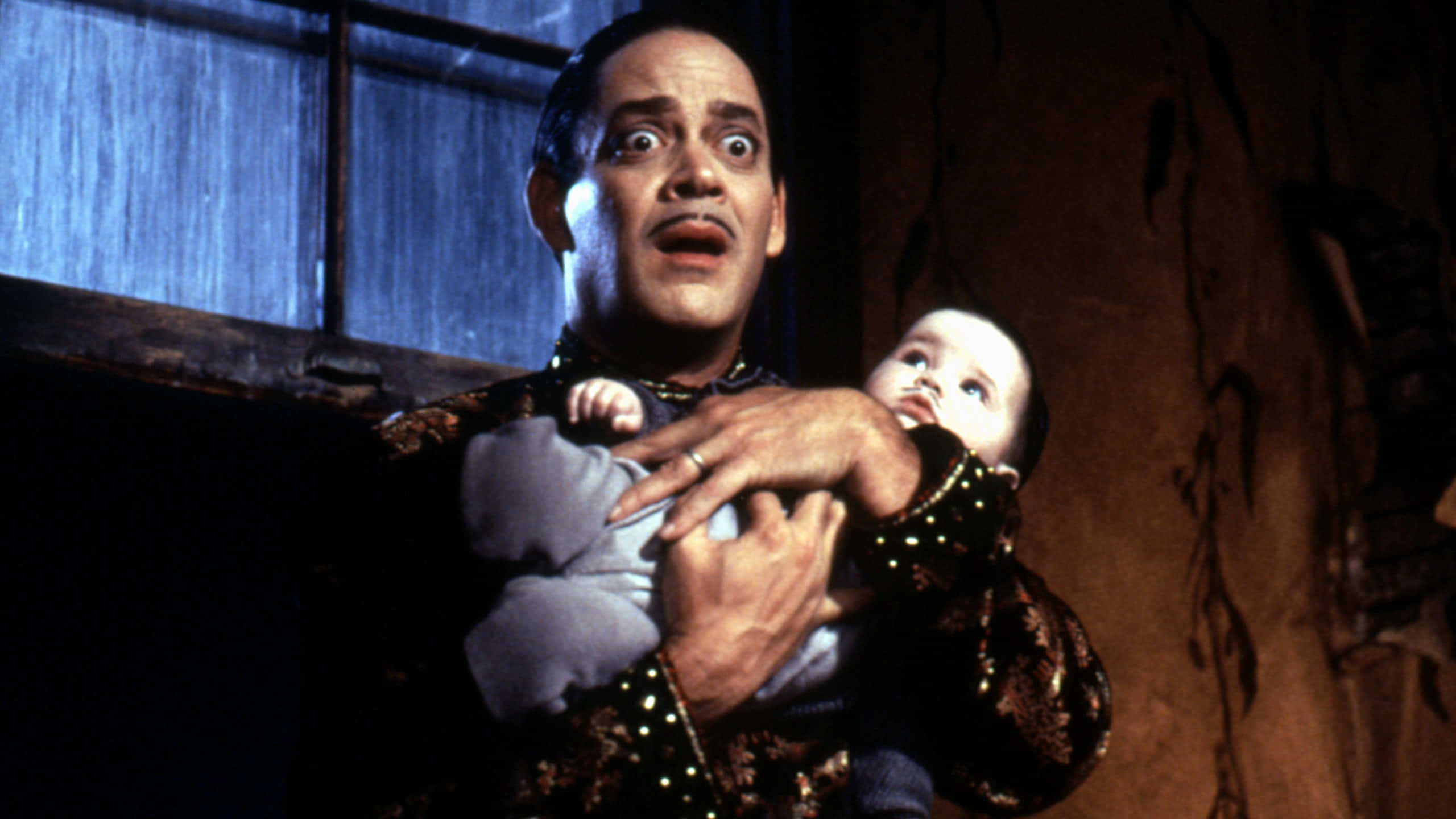 Addams Family 桌布 壁紙 — 一個穿著華麗深色衣服的男人，在一個光線昏暗、窗戶上有雨痕的房間裡，抱著一個睜大眼睛、張開嘴巴的嬰兒。