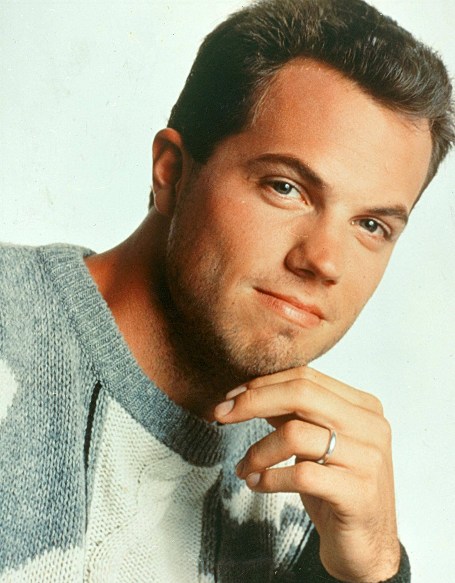 adam baldwin 桌布 壁紙 — 一個穿著藍白針織毛衣的男人，在純色背景前將下巴擱在手上。