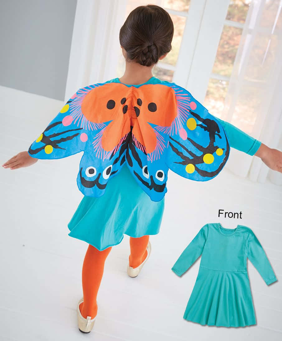 Butterfly Wing Dress 桌布 壁紙 — 一個穿著藍綠色連衣裙的孩子，戴著帶有圓點和眼睛的鮮豔藍色和橙色蝴蝶披肩，站在室內。