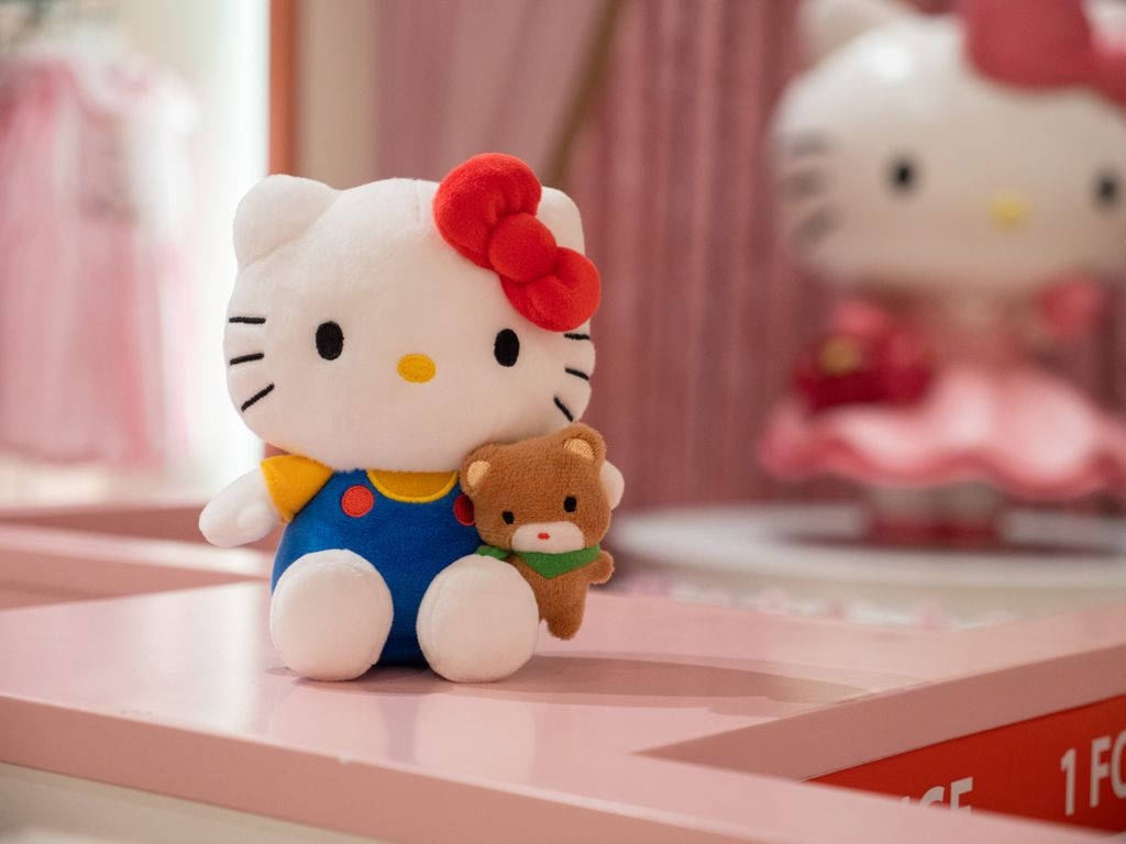 Hello 桌布 壁紙 — 一個穿著藍色吊帶褲的絨毛Hello Kitty在粉紅色架子上抱著一個小泰迪熊，背景是模糊的粉紅色。