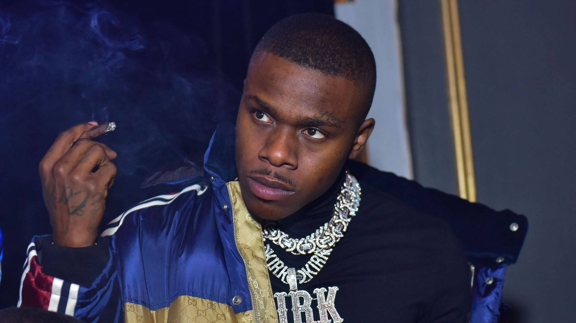 Dababy 桌布 壁紙 — 一個穿著藍色和金色外套的男人，戴著一條粗鑽石鏈，在黑暗背景下抽煙。