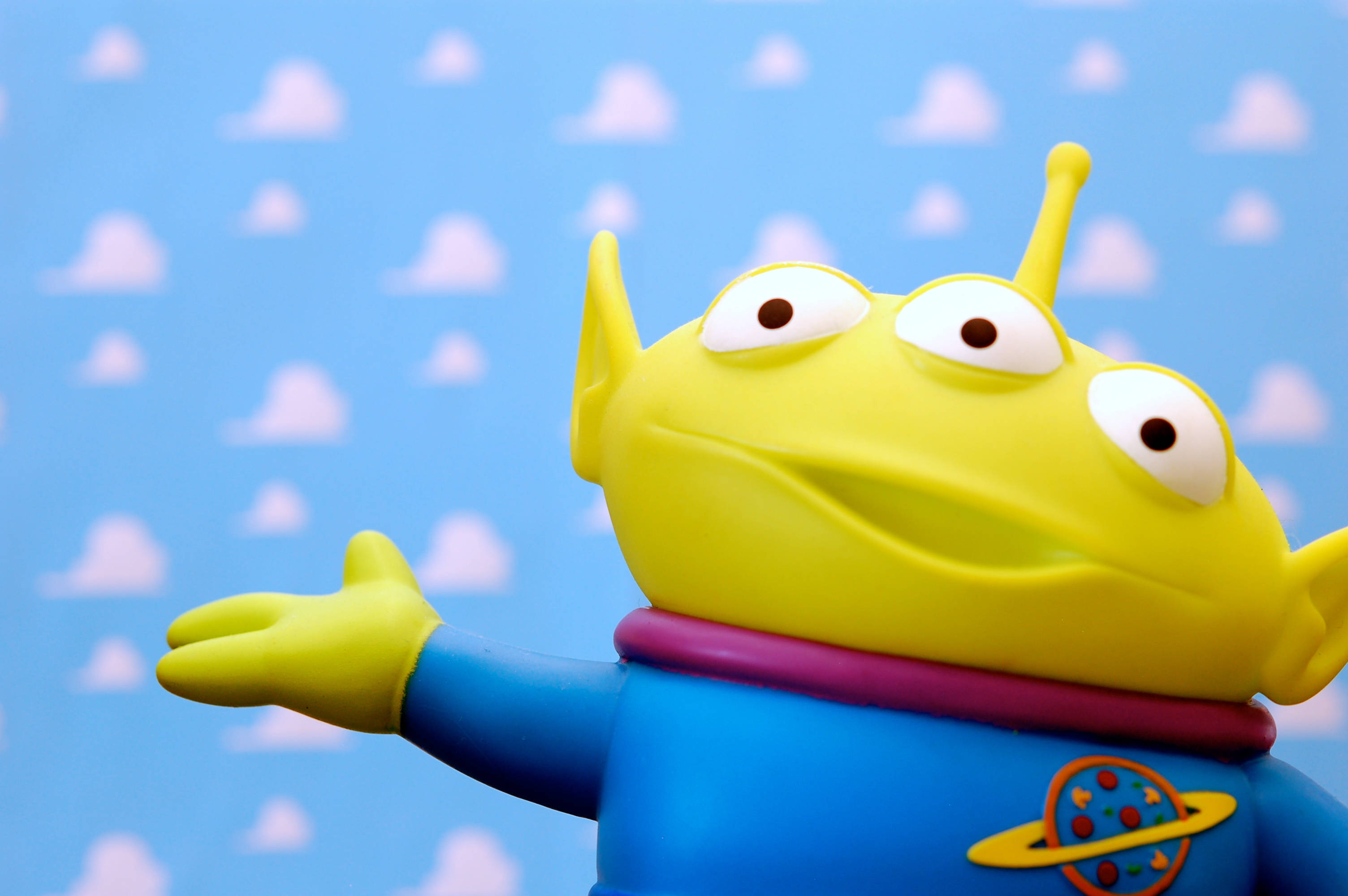 Toy Story Alien 桌布 壁紙 — 一個穿著藍色套裝和紫色領子的快樂黃色三眼外星人玩具，在藍天白雲的背景前揮手。