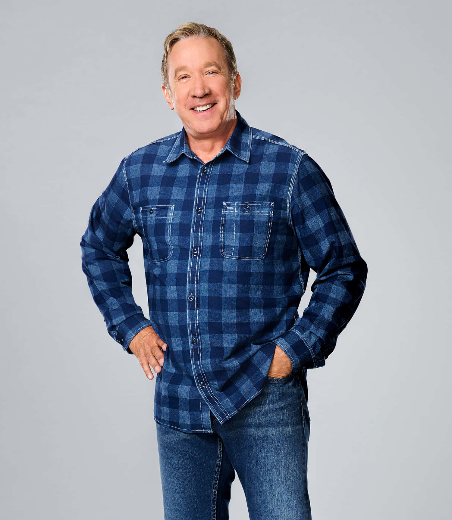 Tim Allen 桌布 壁紙 — 一個穿著藍色格紋襯衫和牛仔褲的開朗男人，雙手插袋，在純灰色背景前微笑。