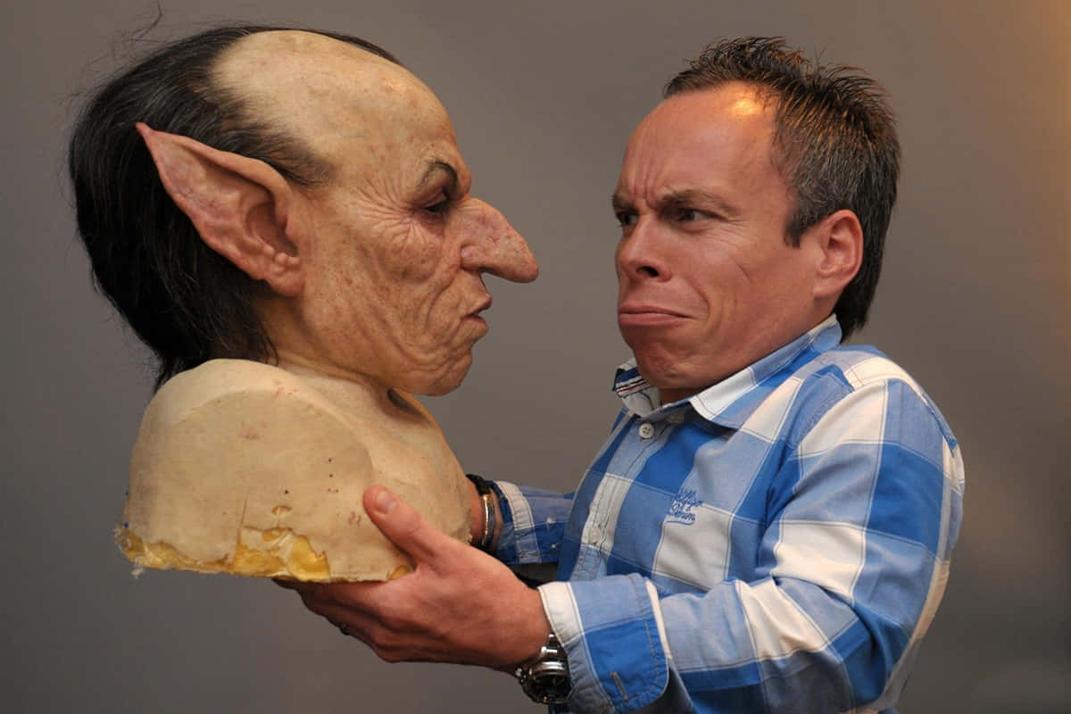 Warwick Davis 桌布 壁紙 — 一個穿著藍色格紋襯衫的男人，在純灰色背景前，拿著一個精緻的哥布林面具，同時模仿它誇張的面部表情。