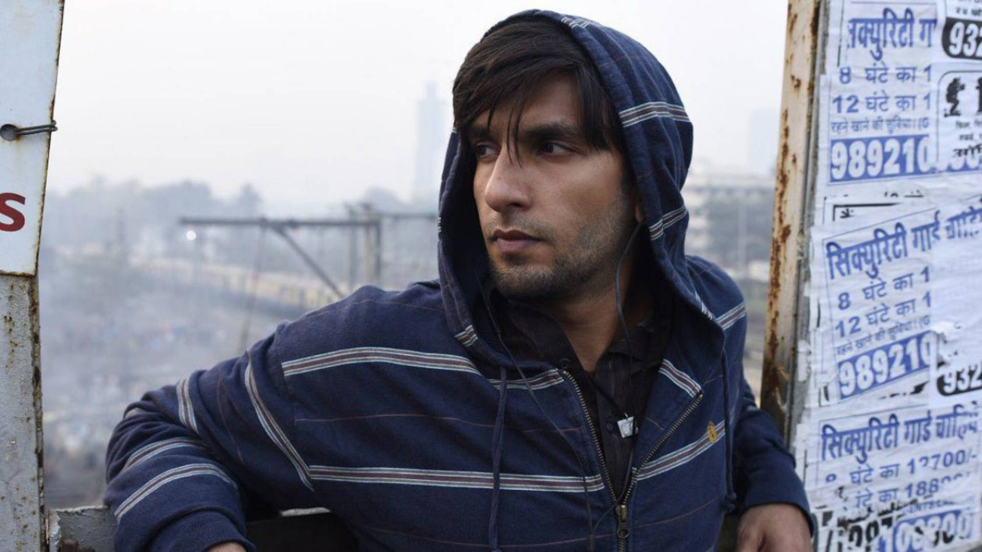 Ranveer Singh 桌布 壁紙 — 一個穿著藍色條紋連帽衫的年輕人，在生鏽的月台上若有所思地凝視著，火車穿過朦朧的城市背景。