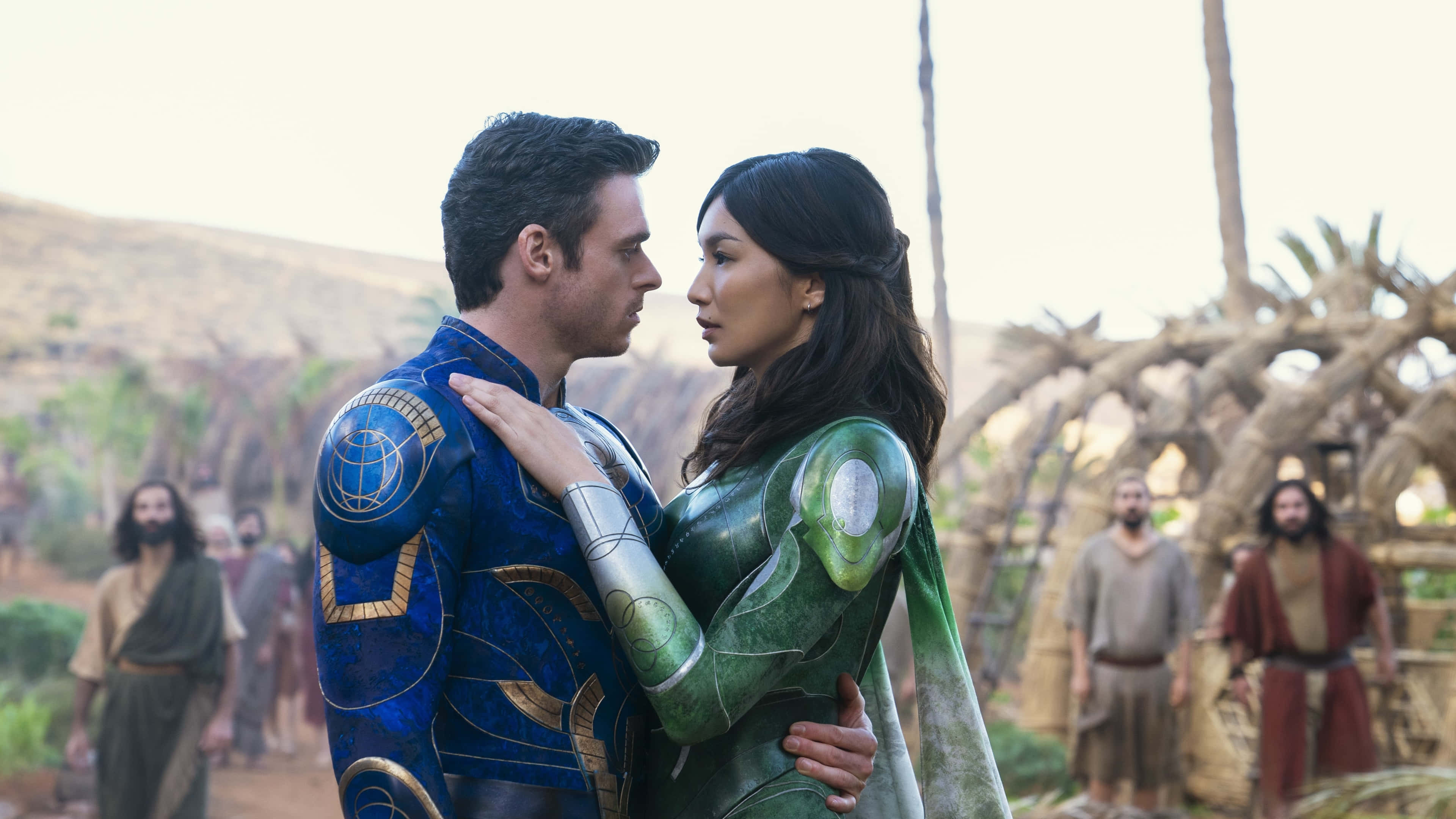 Eternals Movie 桌布 壁紙 — 一個穿著藍色盔甲的男人和一個穿著綠色盔甲的女人在沙漠村莊中與旁觀者分享親密時刻。