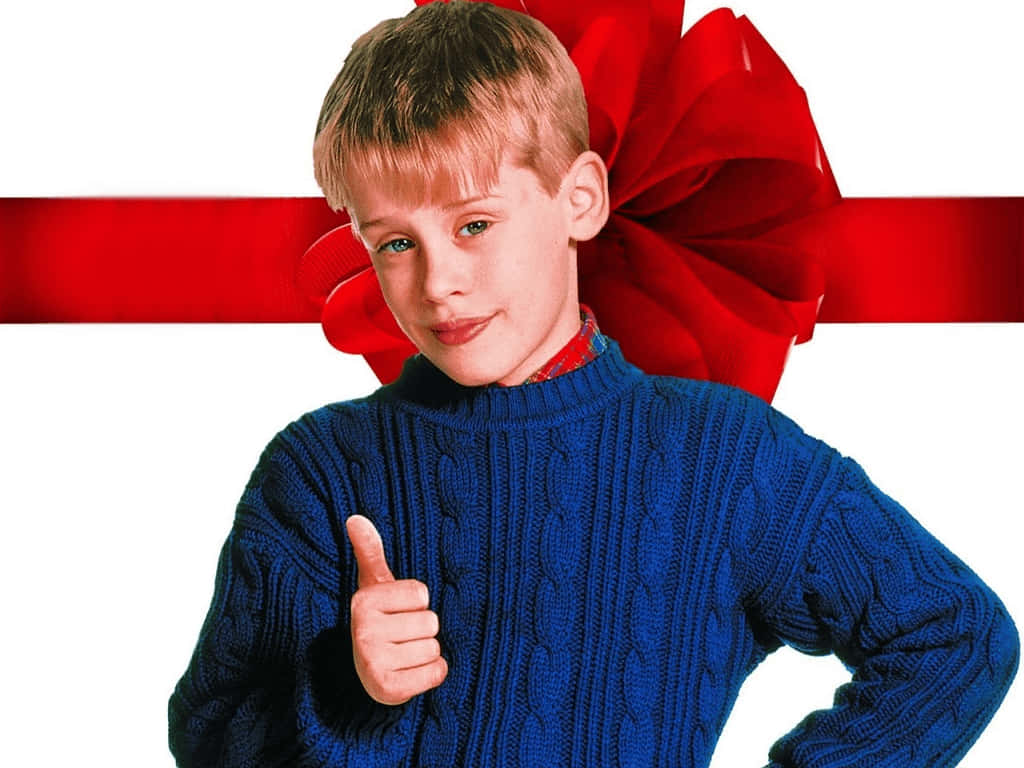 Home Alone 桌布 壁紙 — 一個穿著藍色絞花針織毛衣的小男孩，在白色背景上豎起大拇指，身後有一個大大的紅色蝴蝶結和絲帶。