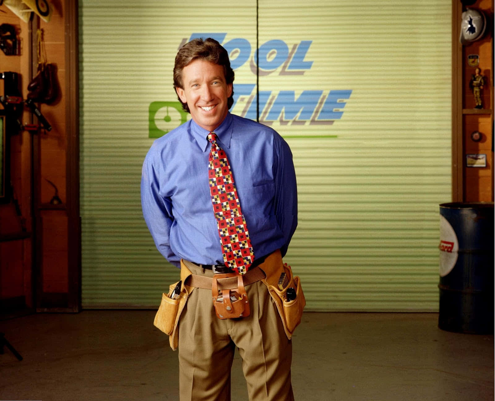 Tim Allen 桌布 壁紙 — 一個穿著藍色襯衫和圖案領帶的開朗男人，站在車庫裡，腰間繫著工具腰帶，身後有「Tool Time」標誌。
