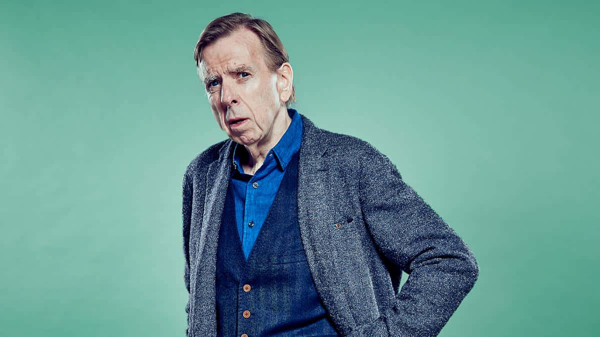 Timothy Spall 桌布 壁紙 — 一個穿著藍色襯衫和灰色西裝外套的男人站在純青色背景前，帶著懷疑的表情。