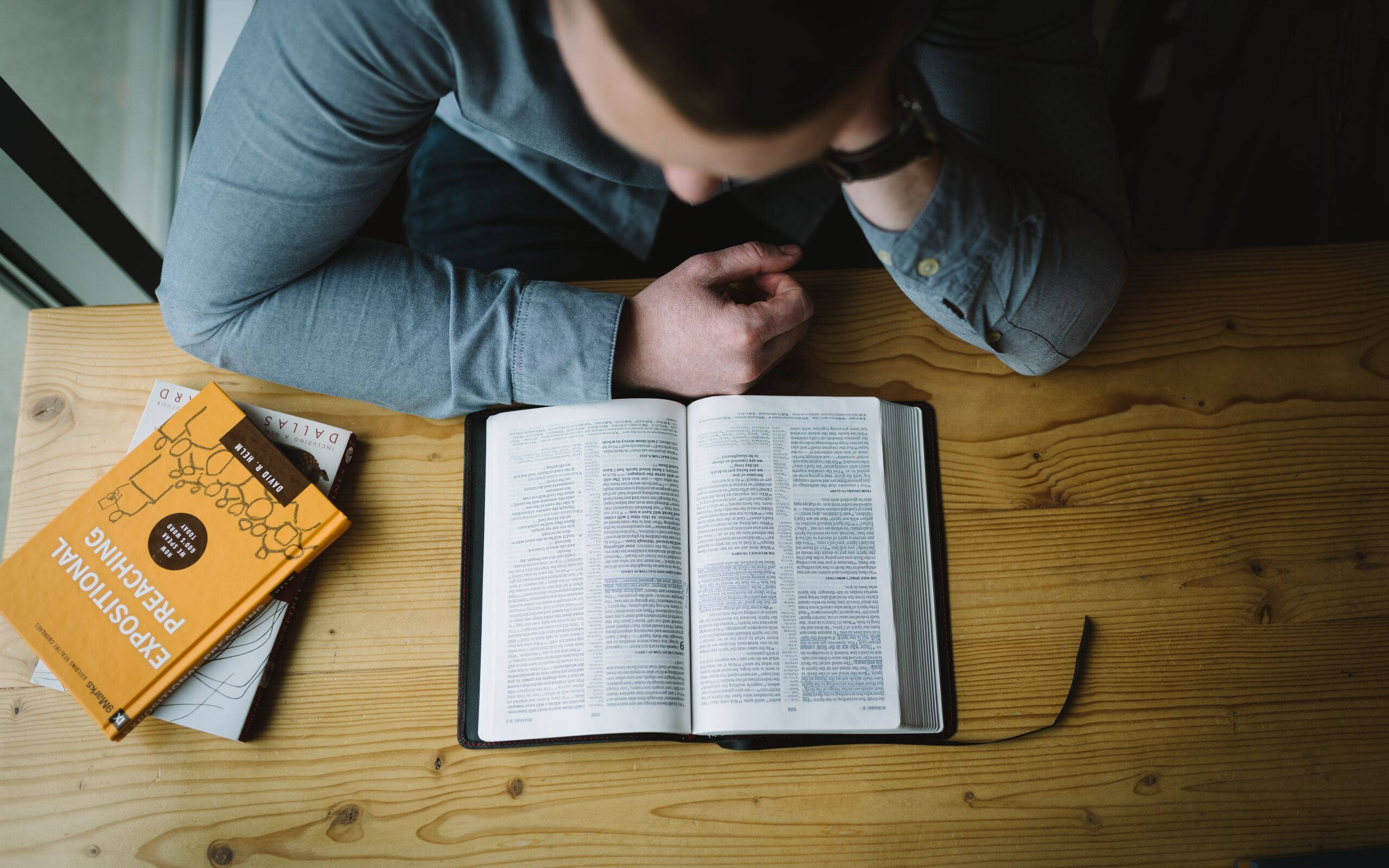 Reading 桌布 壁紙 — 一個穿著藍色襯衫的人俯身在一張木桌上閱讀一本打開的聖經，旁邊有一本黃色的書。