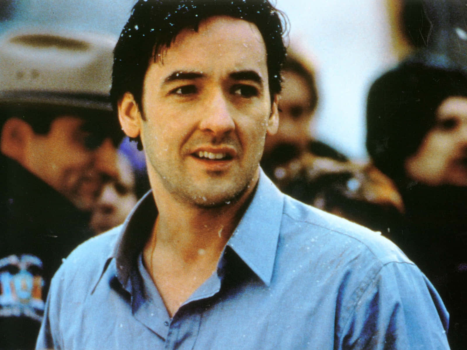 John Cusack 桌布 壁紙 — 一個穿著藍色襯衫的男人在擁擠的戶外場景中微笑，雪花在他周圍飄落。