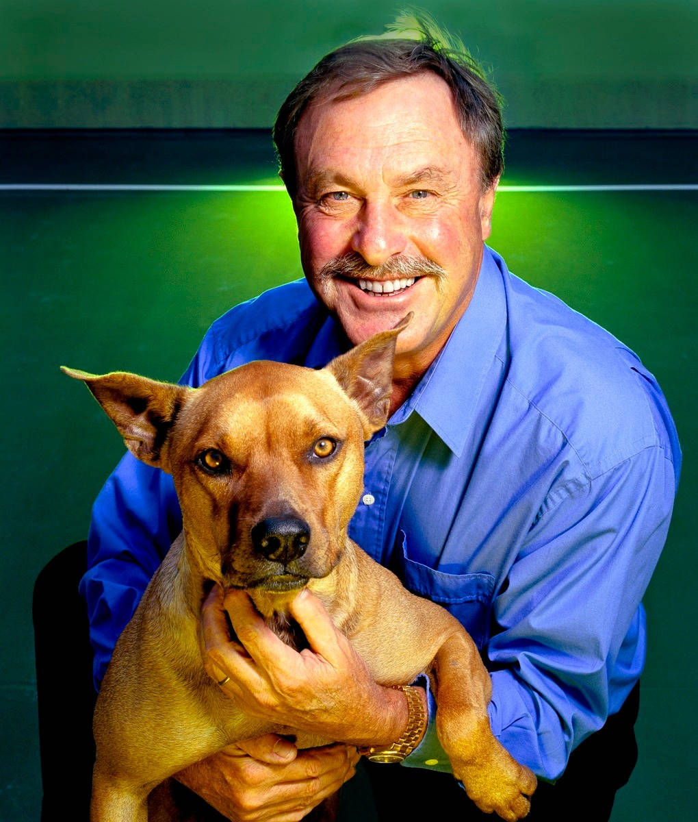 Tennis 桌布 壁紙 — 一個穿著藍色襯衫的開朗男人，在充滿活力的綠色背景前，抱著一隻棕褐色的狗微笑。