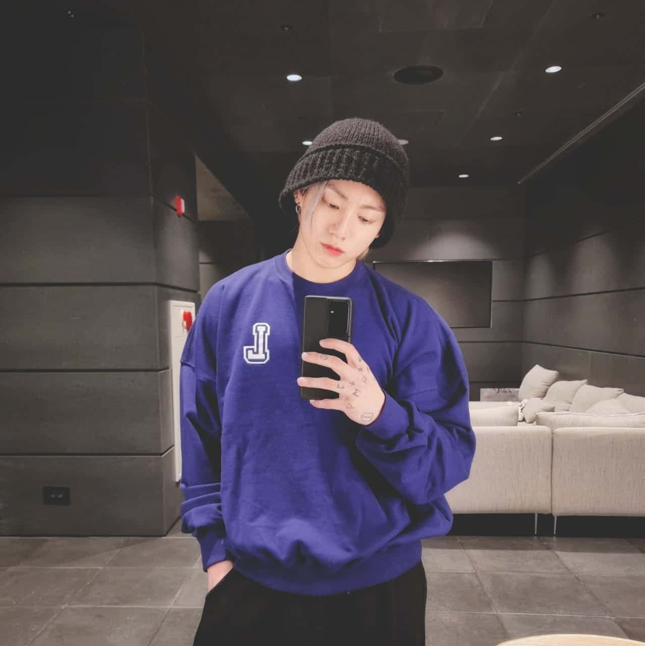 Jungkook 桌布 壁紙 — 一個穿著藍色連帽衫和黑色毛線帽的人，在一個有灰色牆壁和米色沙發的現代簡約房間裡對著鏡子自拍。