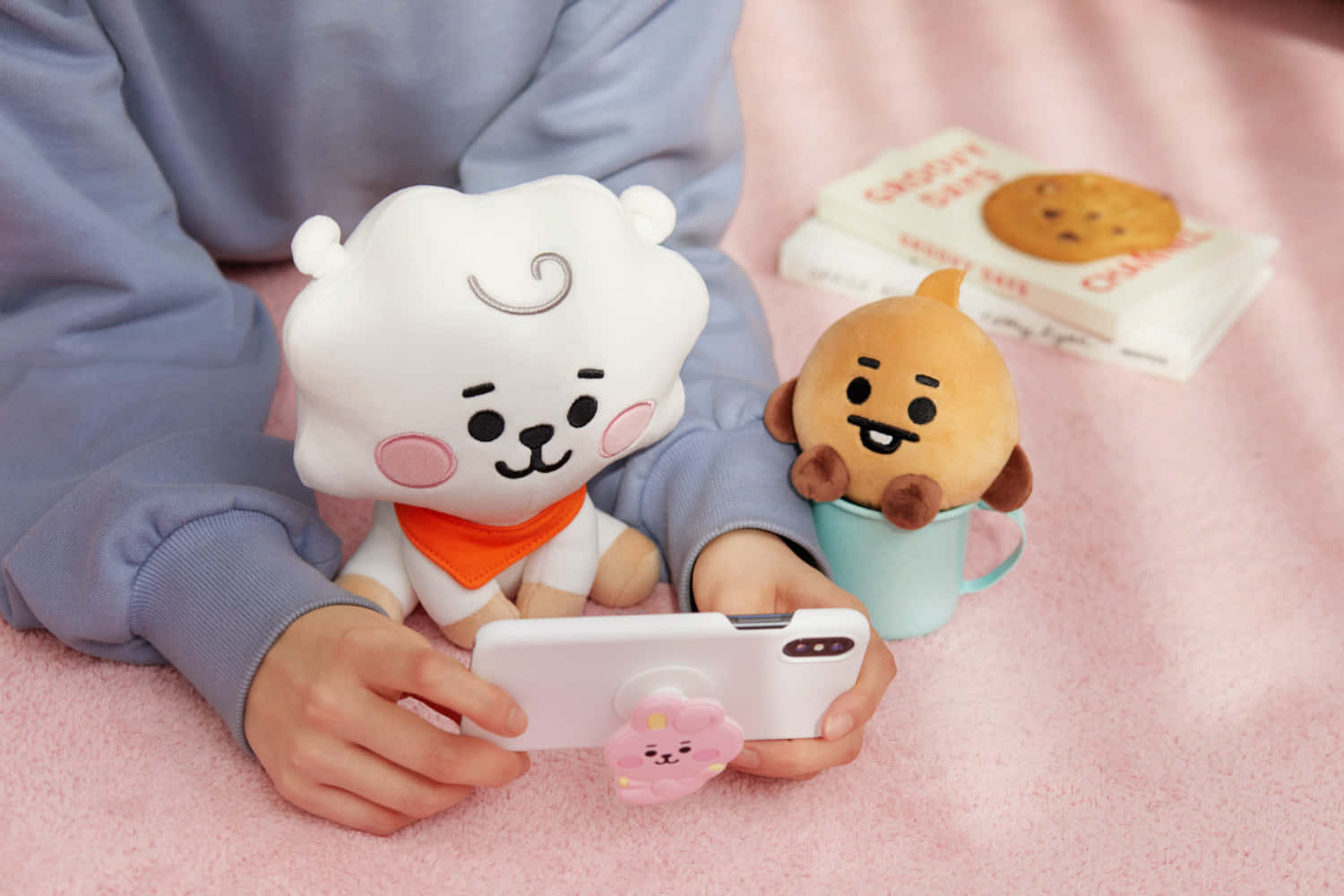 BT21 桌布 壁紙 — 一個穿著藍色連帽衫的人拿著一部帶有粉紅色角色握把的手機，旁邊有兩個BT21毛絨玩具坐在粉紅色毯子上。