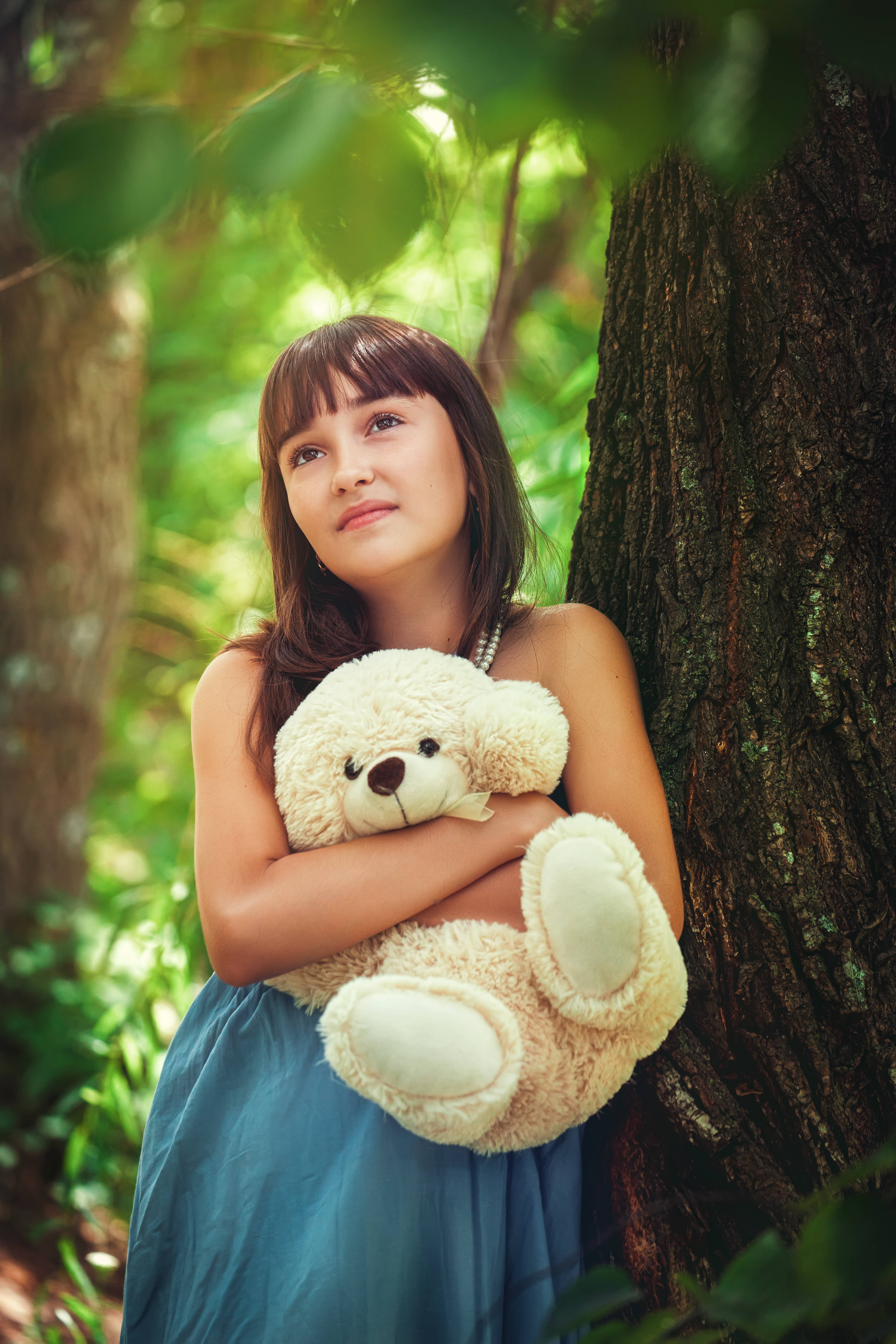 Cute Teddy Bear 桌布 壁紙 — 一個穿著藍色連衣裙的小女孩在陽光明媚的森林裡靠著樹，抱著一個毛絨絨的米色泰迪熊。