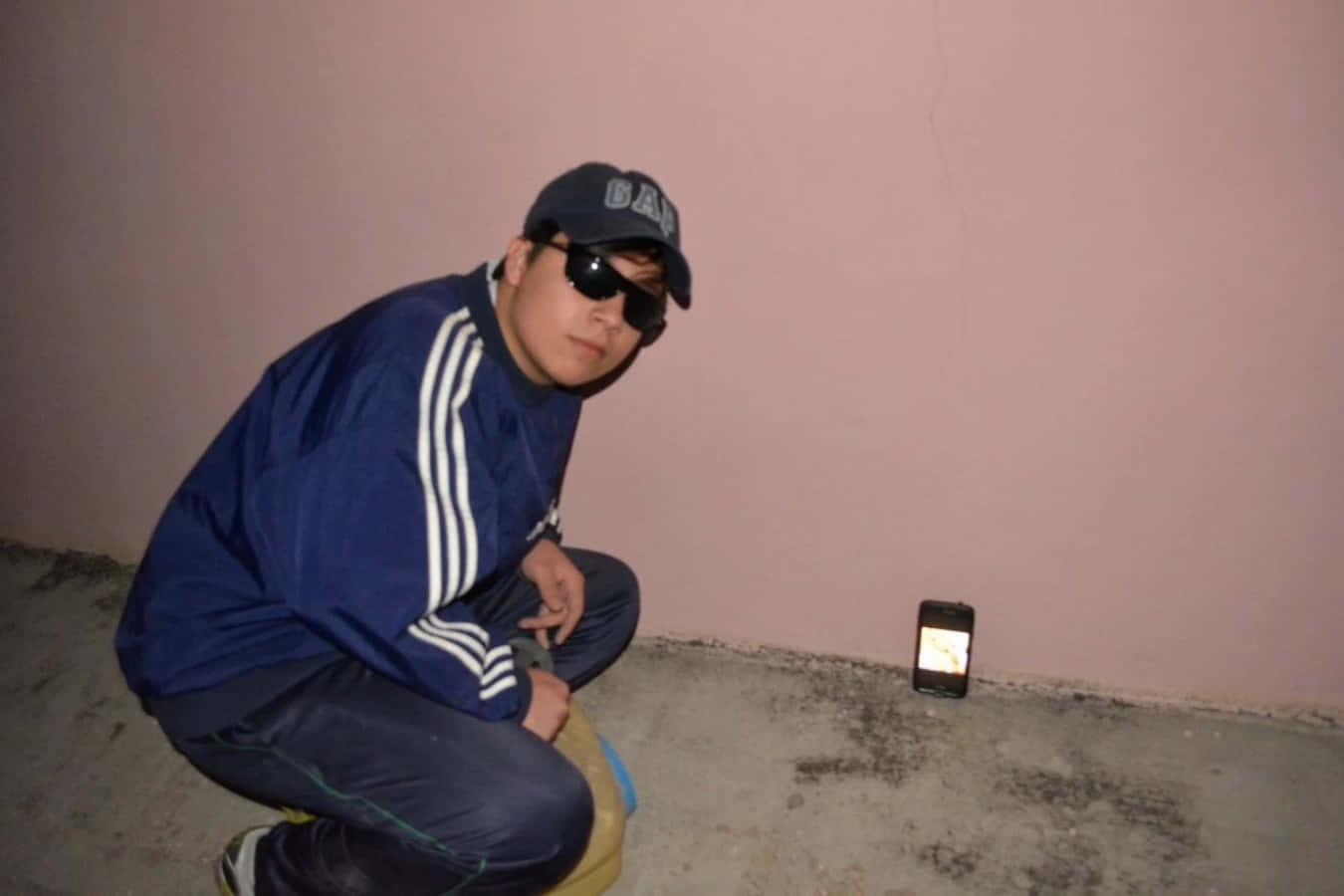 Quackity 桌布 壁紙 — 一個穿著藍色運動服和帽子的人蹲在發光的手機旁邊，背景是灰塵粉紅色的牆壁。