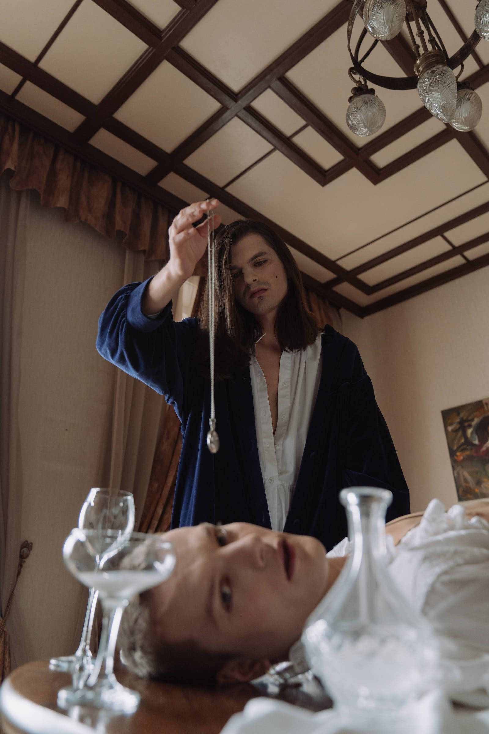 Hypnosis 桌布 壁紙 — 一個穿著藍色長袍的人，在一個光線昏暗、有吊燈的房間裡，將一個鐘擺懸在躺在桌子上方的另一個人上方。