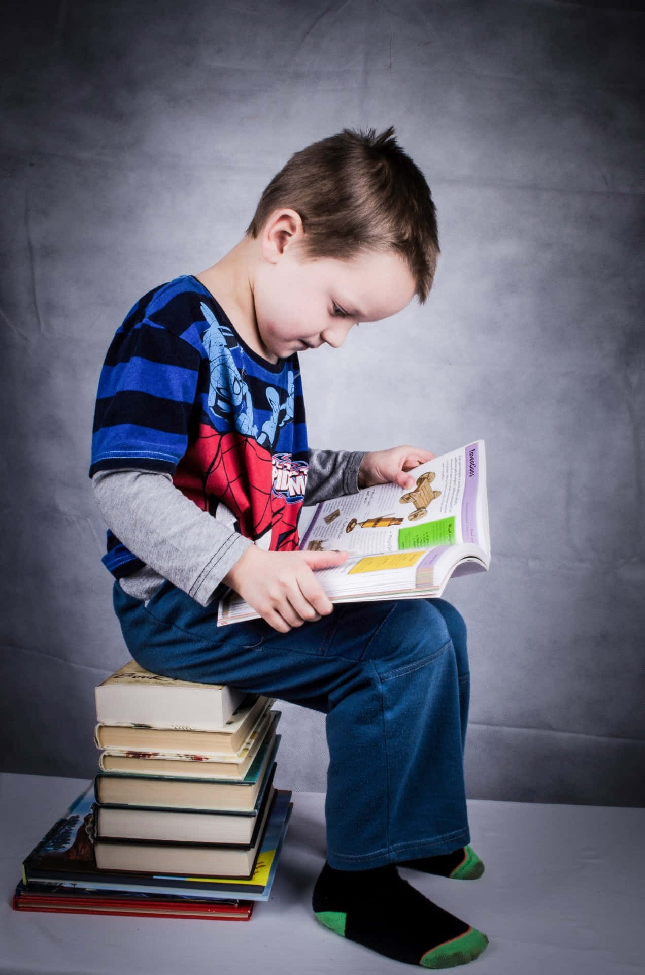 Boy 桌布 壁紙 — 一個穿著蜘蛛人T恤的小男孩坐在一堆書上，在灰色工作室背景下專心閱讀。