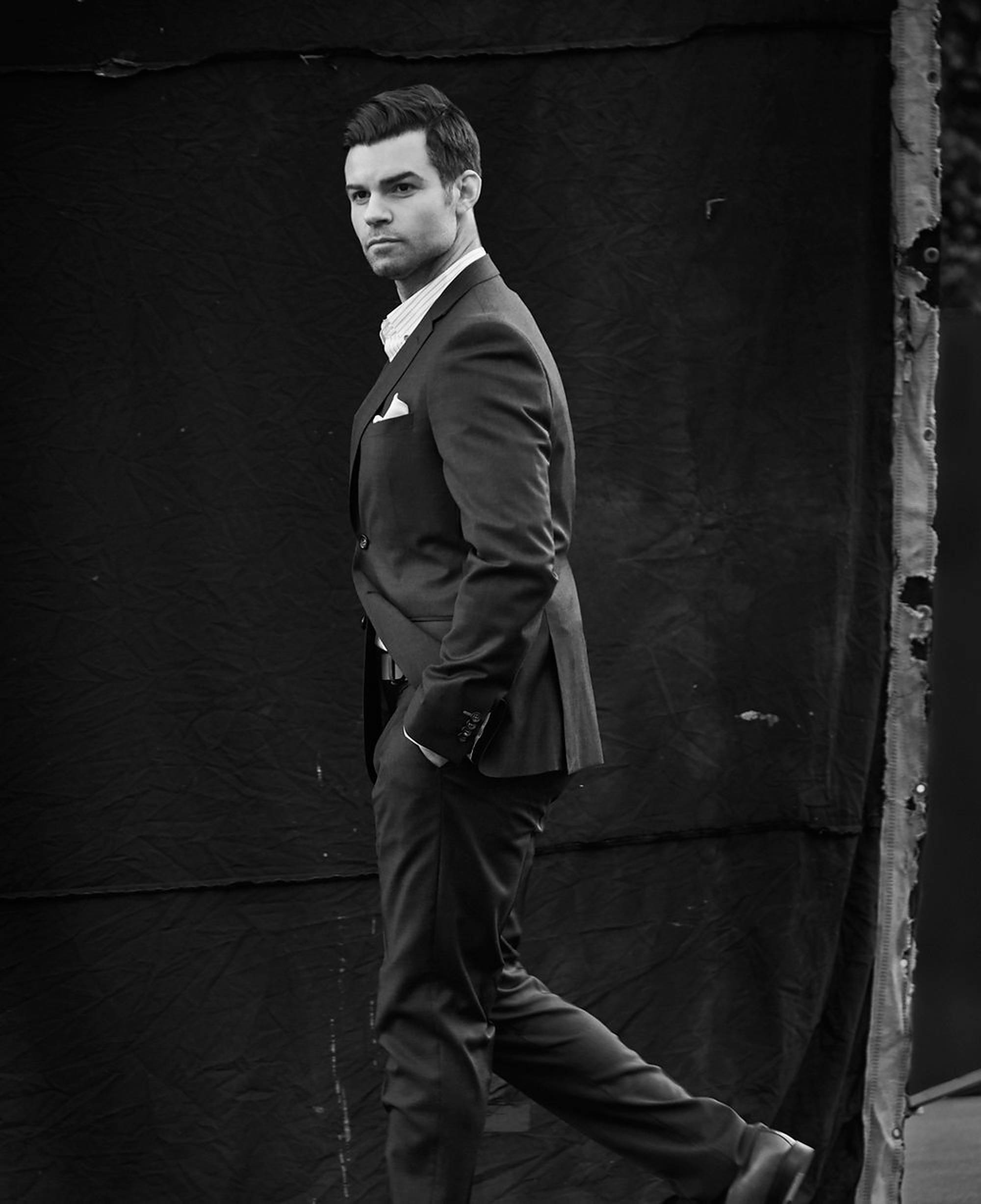 Daniel Gillies 桌布 壁紙 — 一個穿著西裝、打扮俐落的男士，在帶有紋理的黑色背景前自信地大步走，形成一幅黑白肖像。