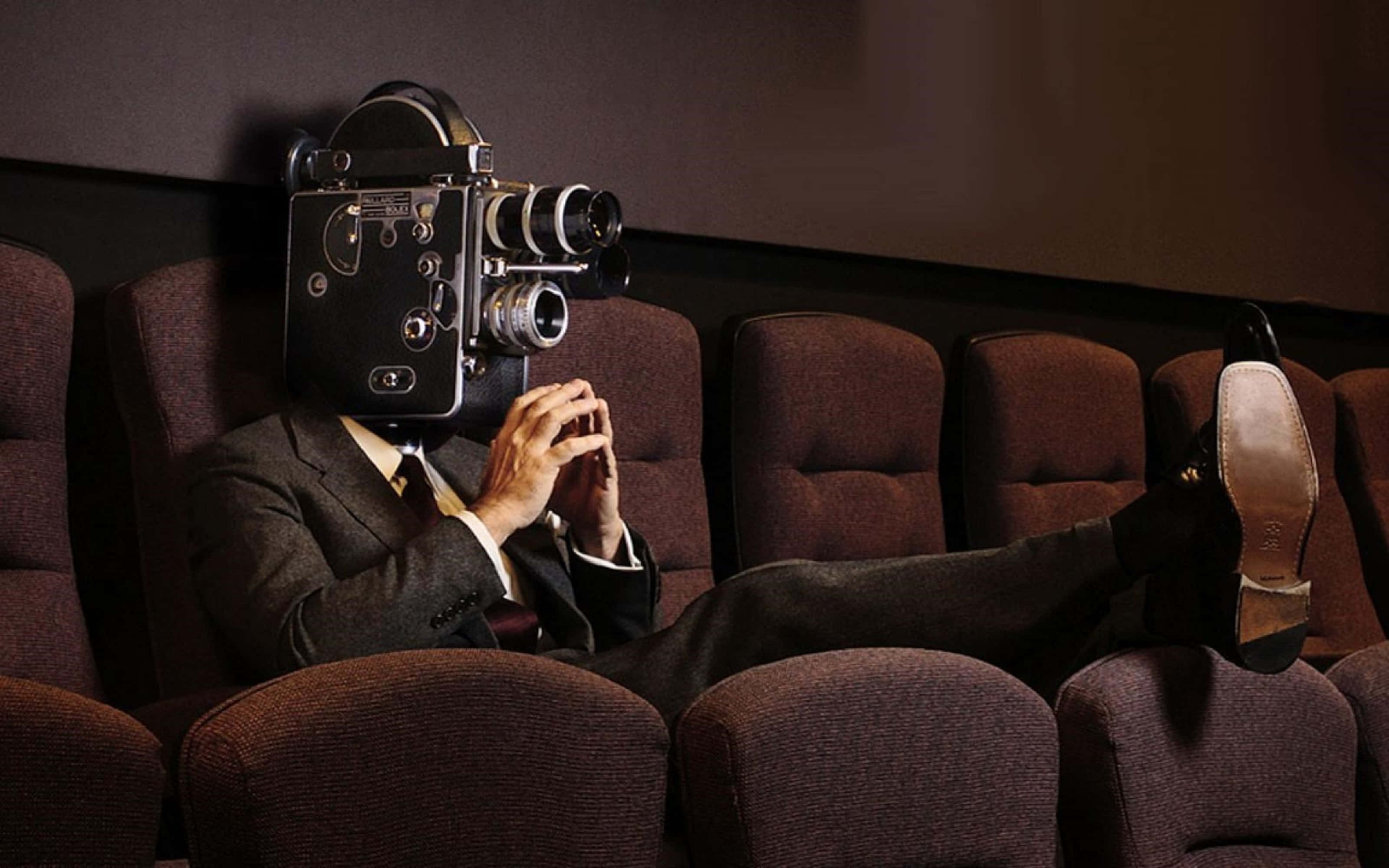 4K Projector 桌布 壁紙 — 一個穿著西裝、頭部是復古電影攝影機的男人，放鬆地坐在昏暗的戲院座位上，雙腳翹起，正在看電影。
