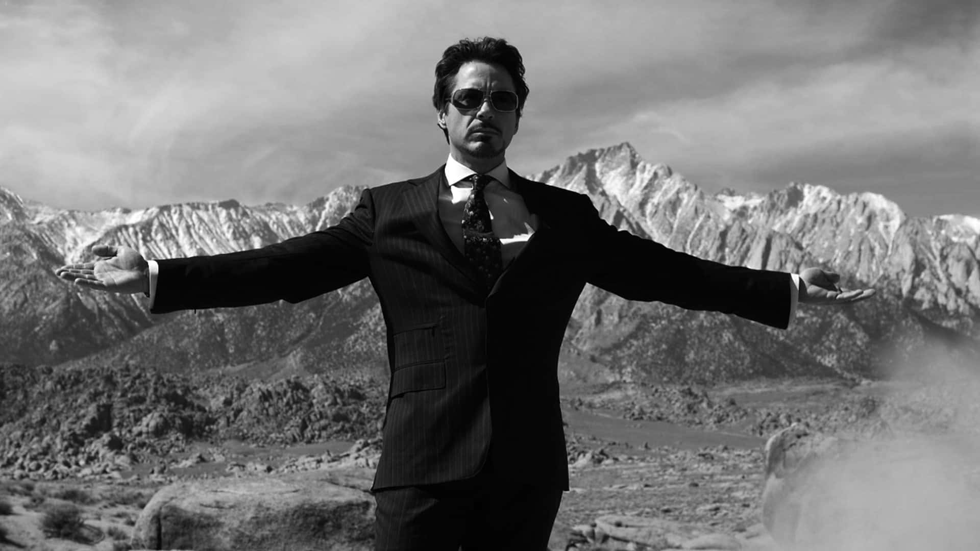 Iron Man Black And White 桌布 壁紙 — 一個穿著西裝和太陽眼鏡的男人在黑白荒涼的山景中張開雙臂站立。