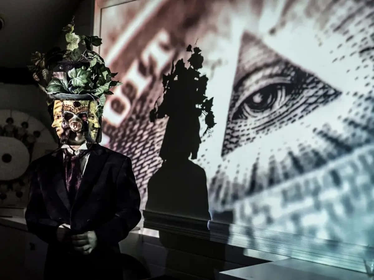 illuminati 桌布 壁紙 — 一個穿著西裝的人物戴著由綠色藤蔓和面孔組成的頭飾，站在投射的眼睛特寫和文字前。