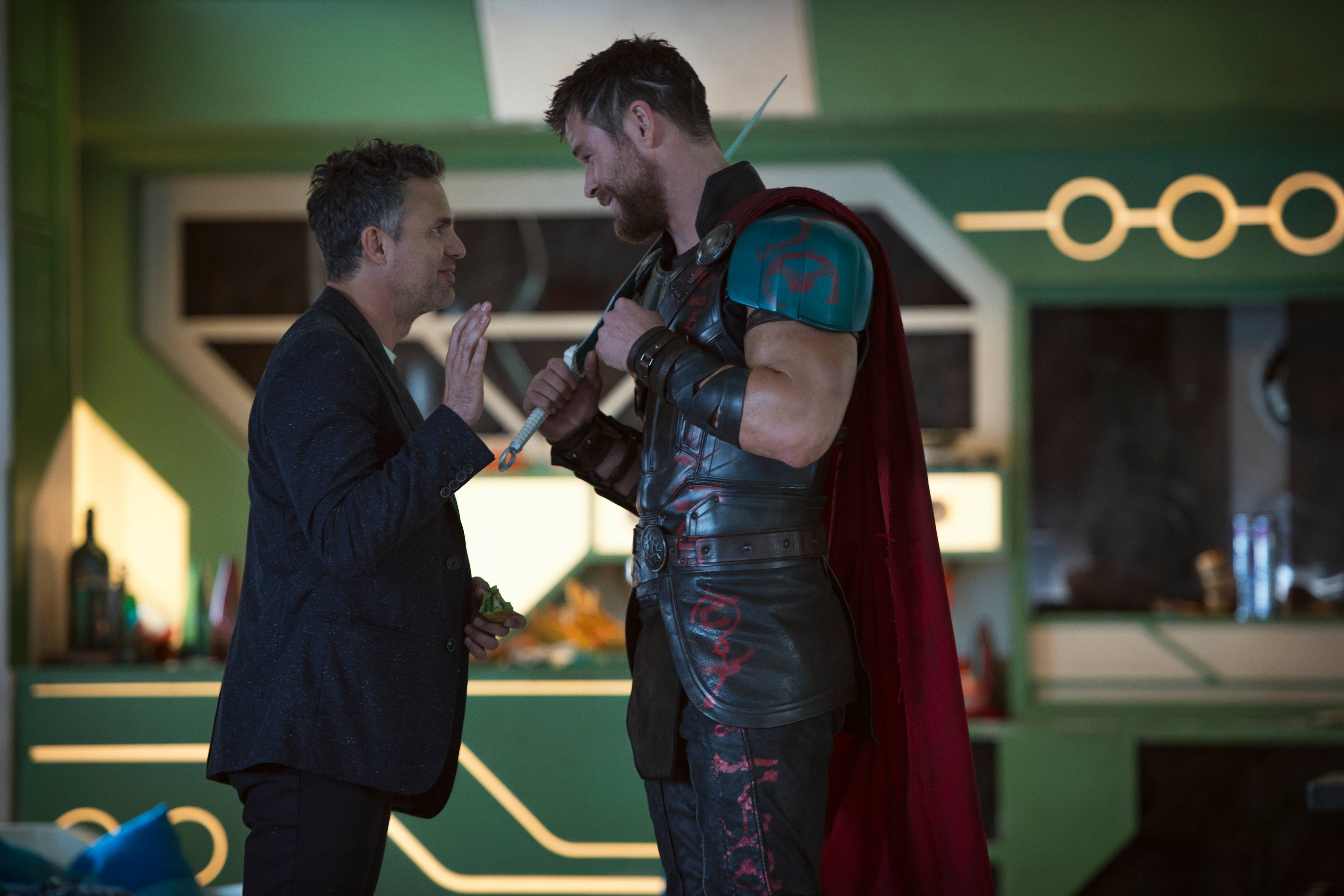 Mark Ruffalo 桌布 壁紙 — 一個穿著西裝的男人在一個未來風格的綠色和金色房間內向穿著戰鬥盔甲的索爾做手勢。