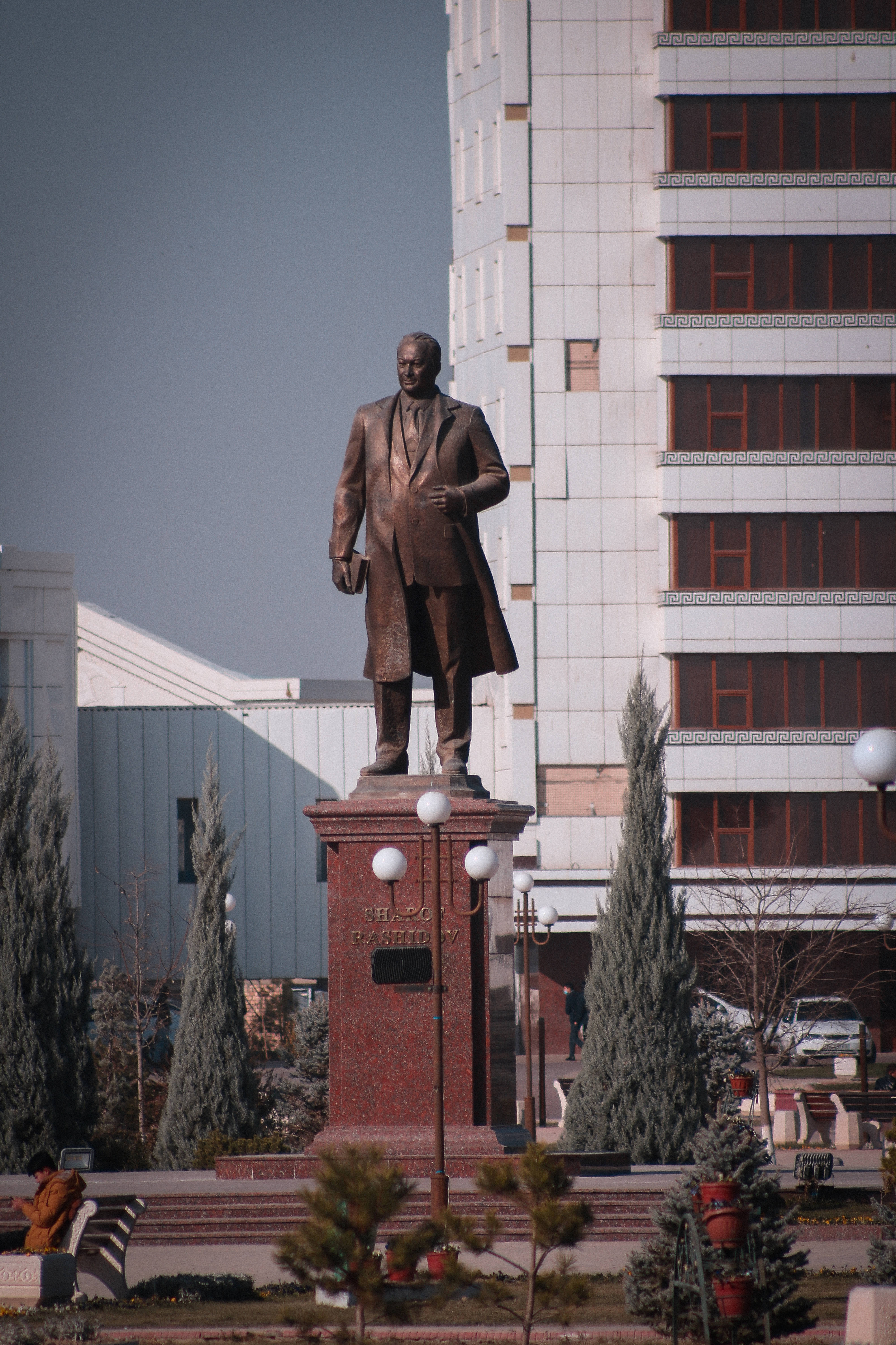 Uzbekistan 桌布 壁紙 — 一個穿著西裝的男人的銅像高高矗立在城市廣場的紅色基座上，周圍是現代建築和常綠樹木，天空晴朗。