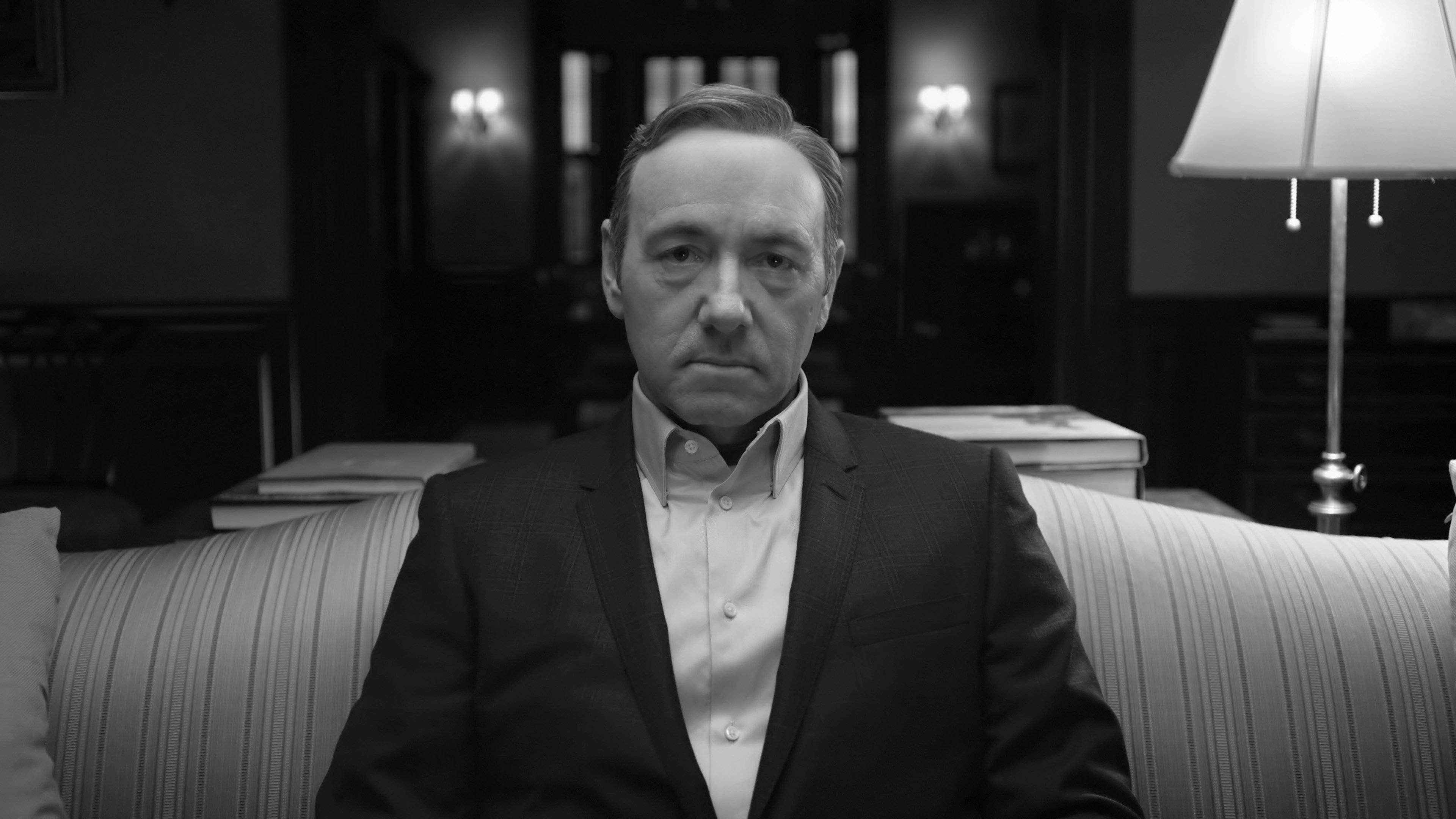house of cards 桌布 壁紙 — 一個穿著西裝的男人莊嚴地坐在光線昏暗的房間裡的條紋沙發上，身後有一盞燈投射出柔和的光線。